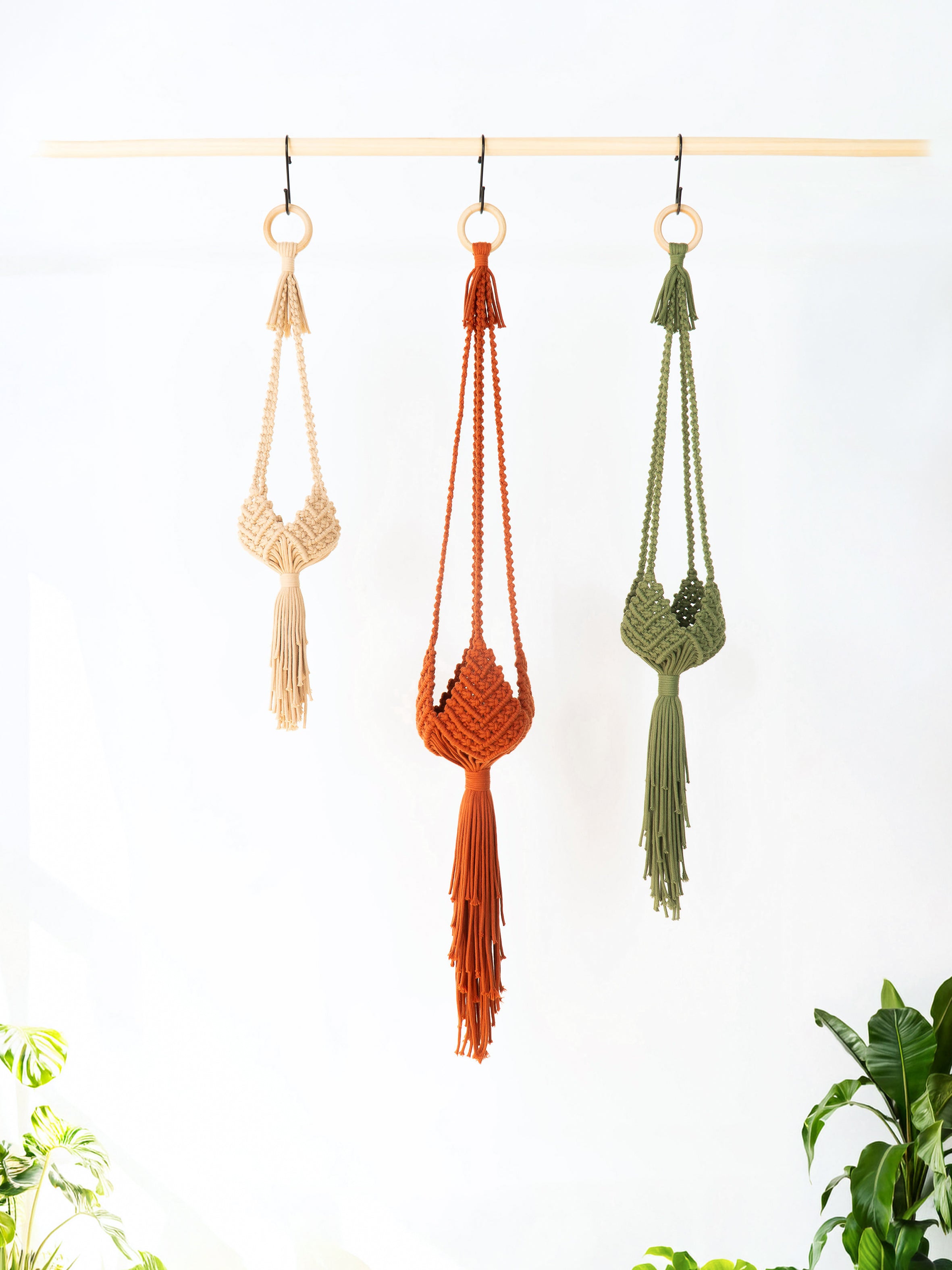 Three macrame wall hangings in beige, rust orange, and sage green hang from a light wood rod against a white wall, with small green plants visible at the bottom corners.