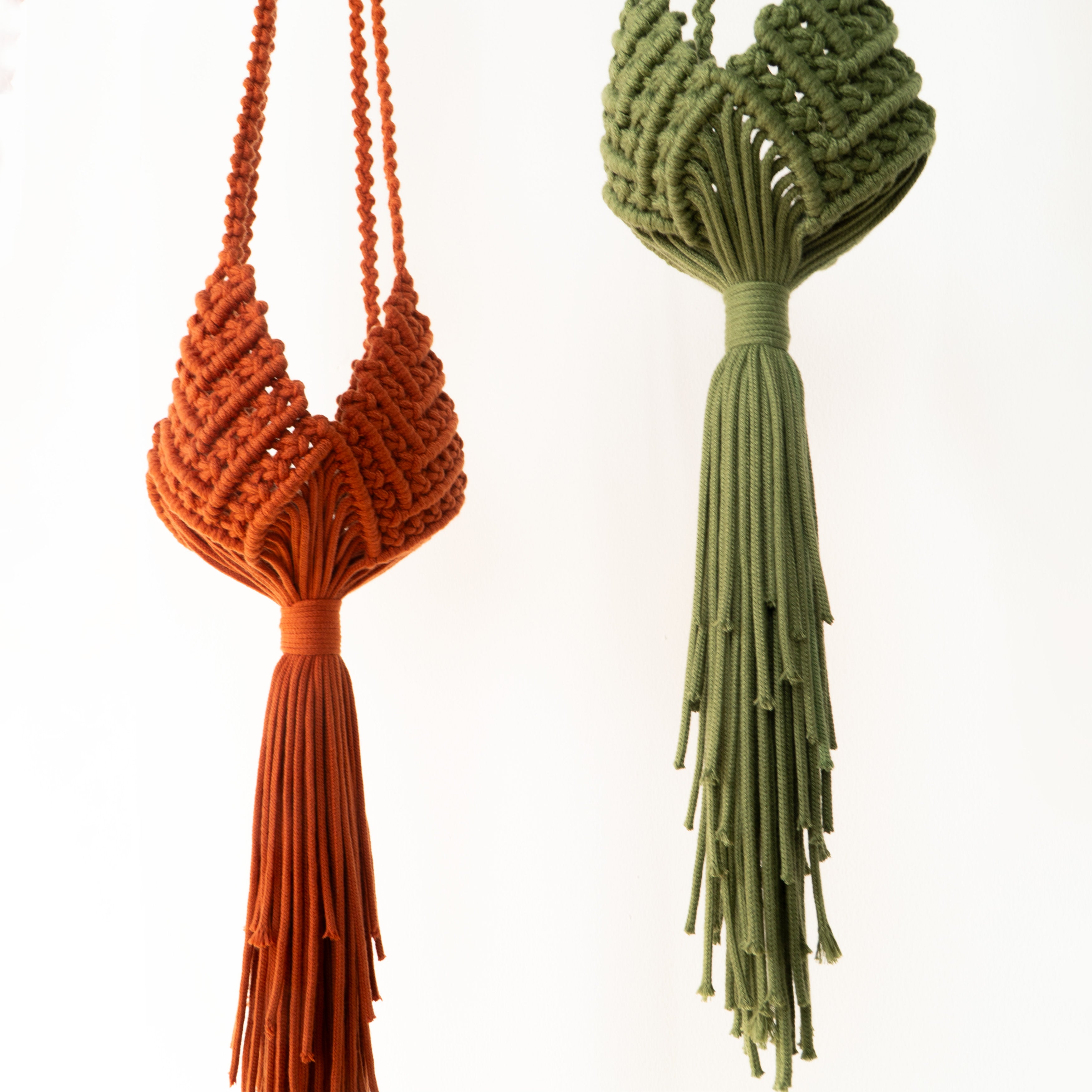 Two macrame wall hangings shown against a plain white background: one in burnt orange with a long tassel, and one in olive green with a frayed tassel.
