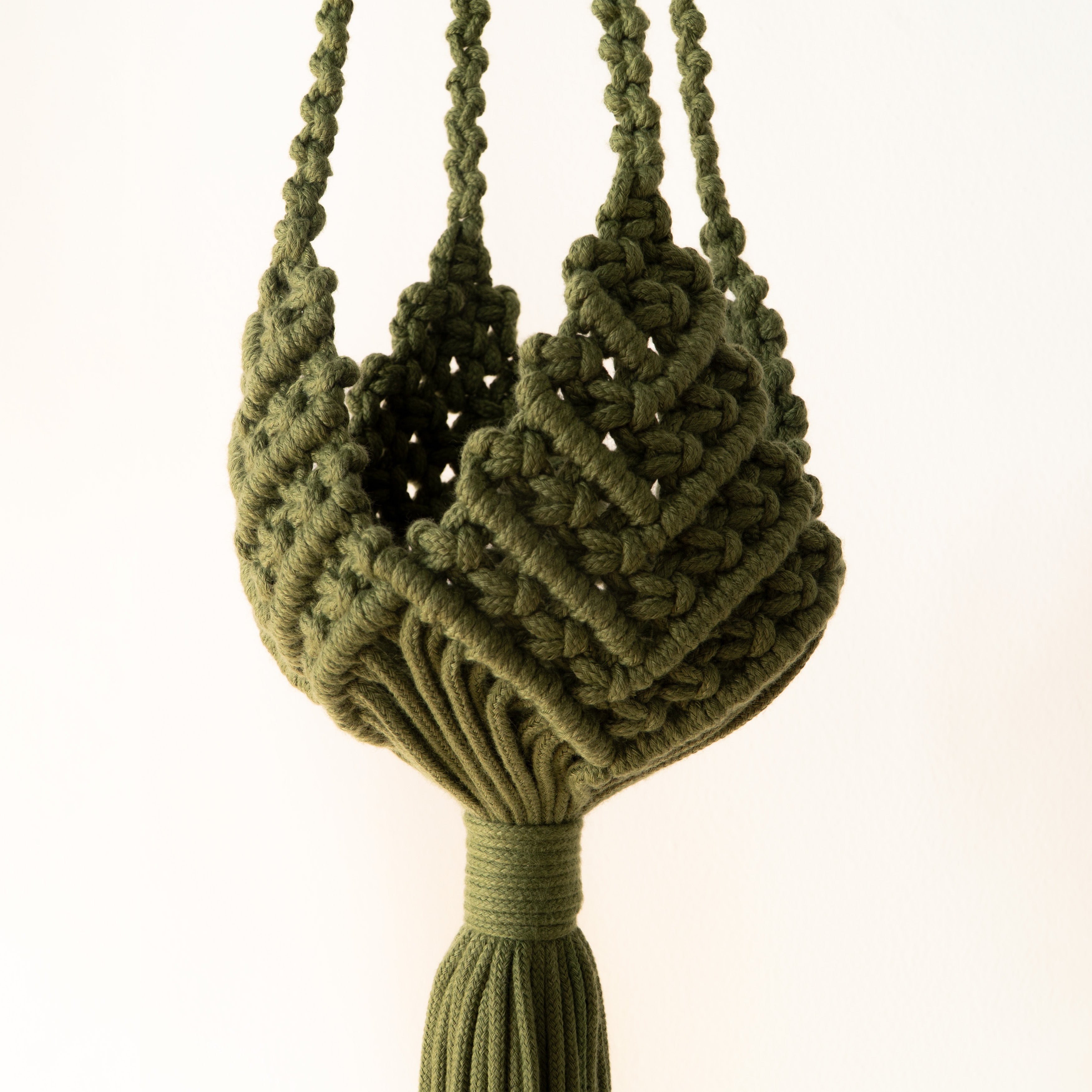 An olive green macrame plant hanger with a textured woven body and a tassel at the bottom, suspended against a plain white background.