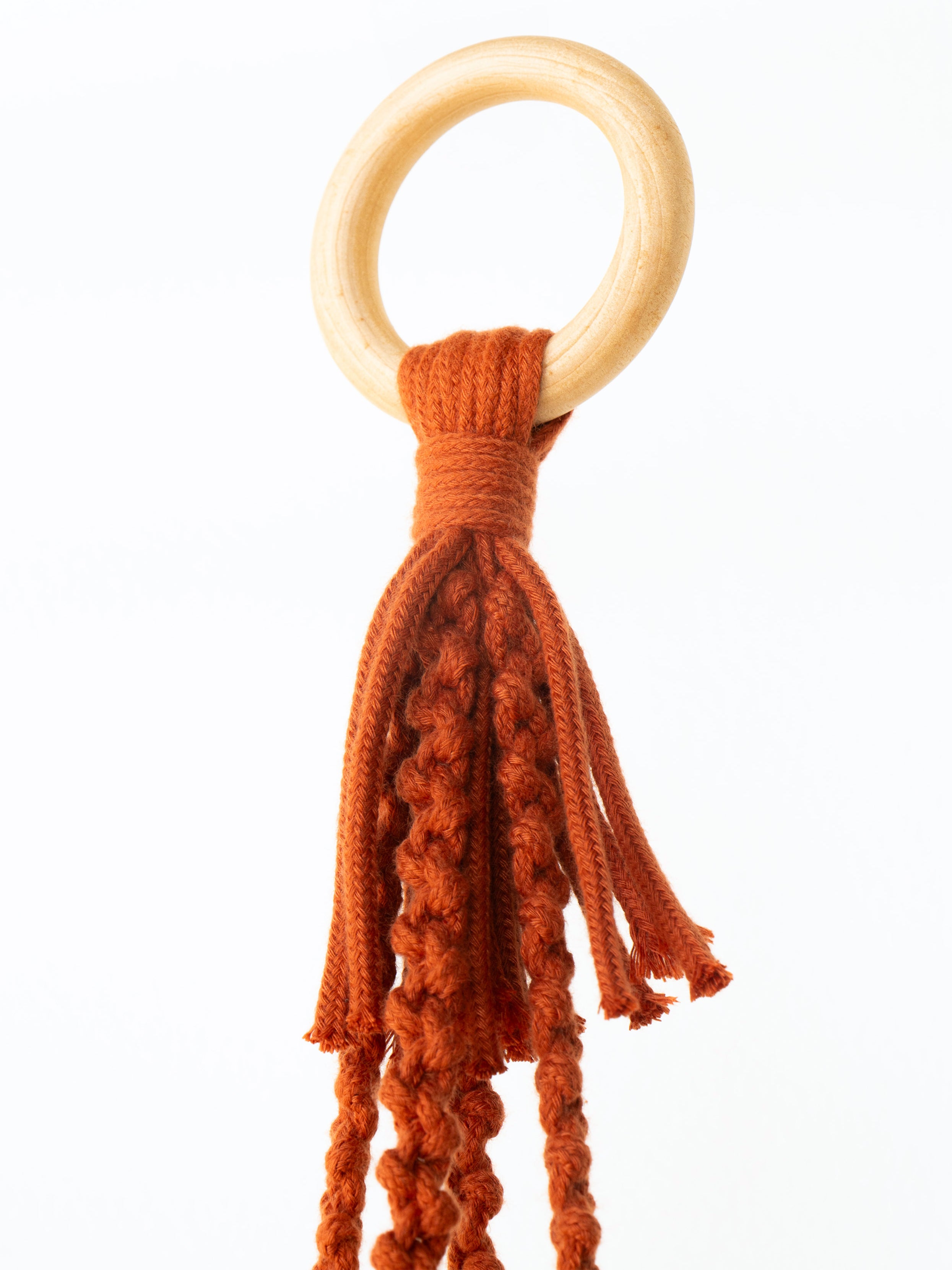 A baby teething ring toy featuring a natural light wood O-ring attached to a rust-orange knotted cotton tassel, shown against a plain white background.