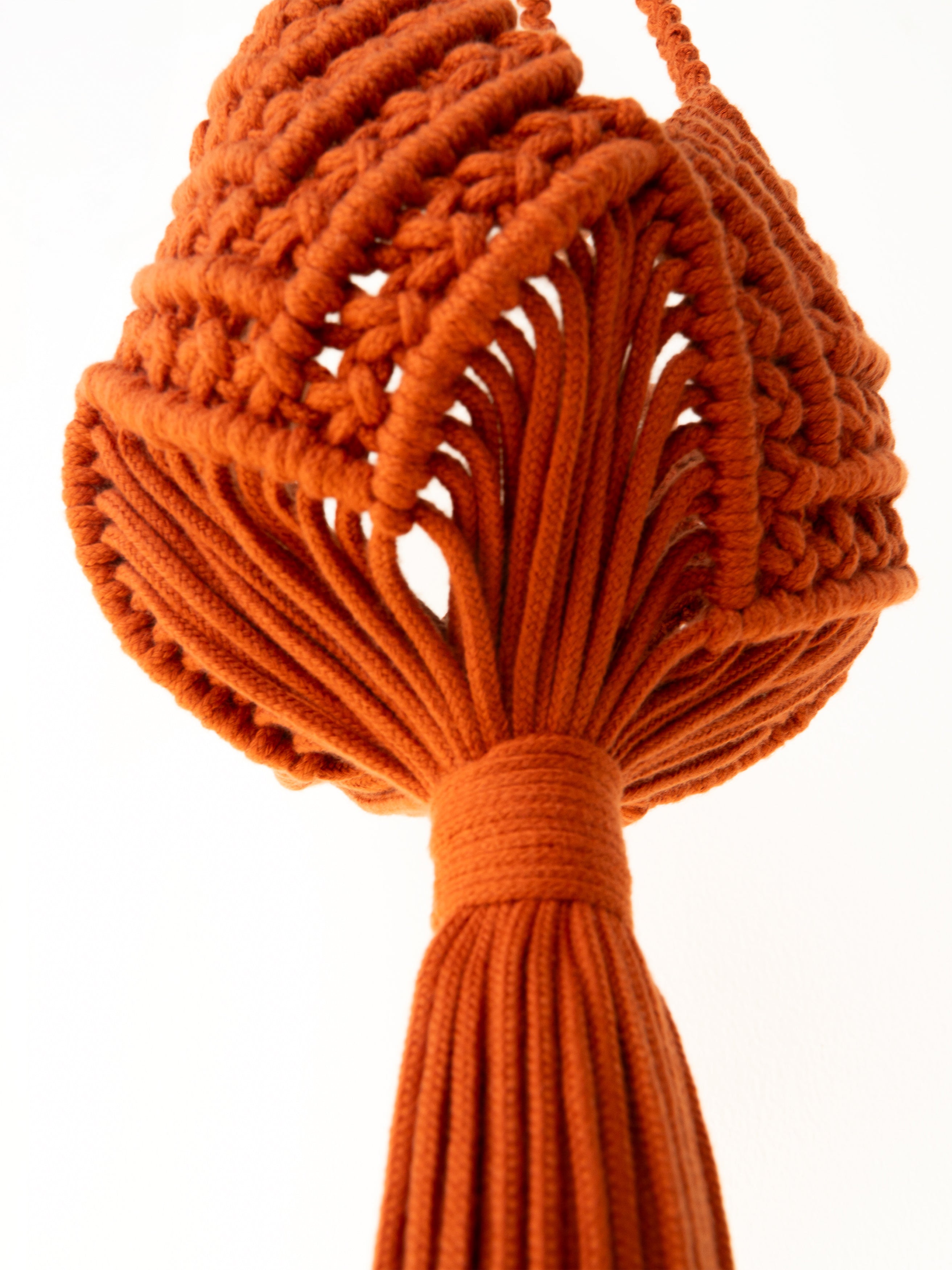 An orange macrame wall hanging with a textured woven top and a long tassel, shown against a plain white background.