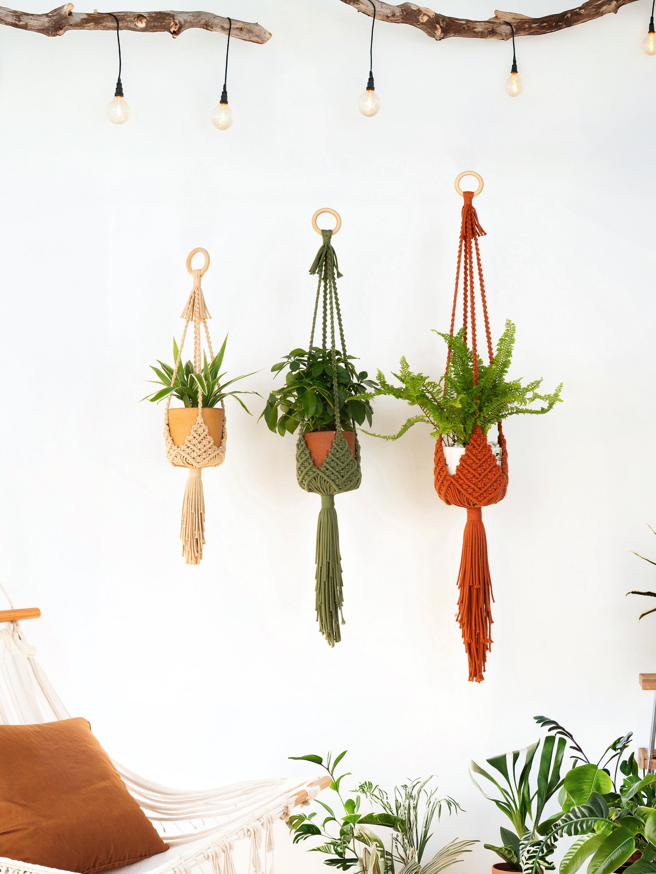 Three macrame plant hangers in beige, green, and rust orange hang on a white wall in a styled room setting with string lights, a hammock, and potted plants.