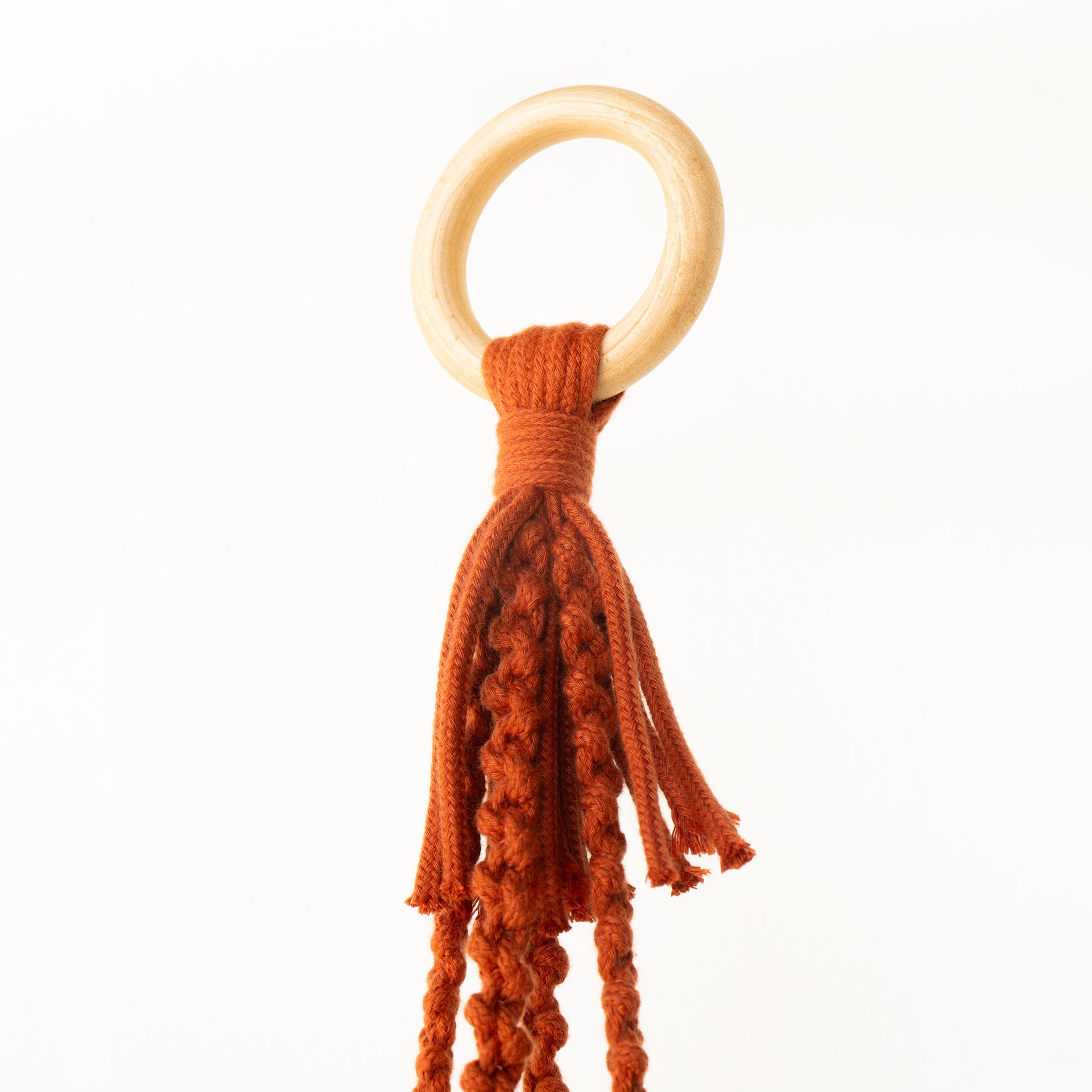 A baby teething ring toy featuring a natural light wood O-ring attached to a rust-orange macrame tassel, shown against a plain white background.