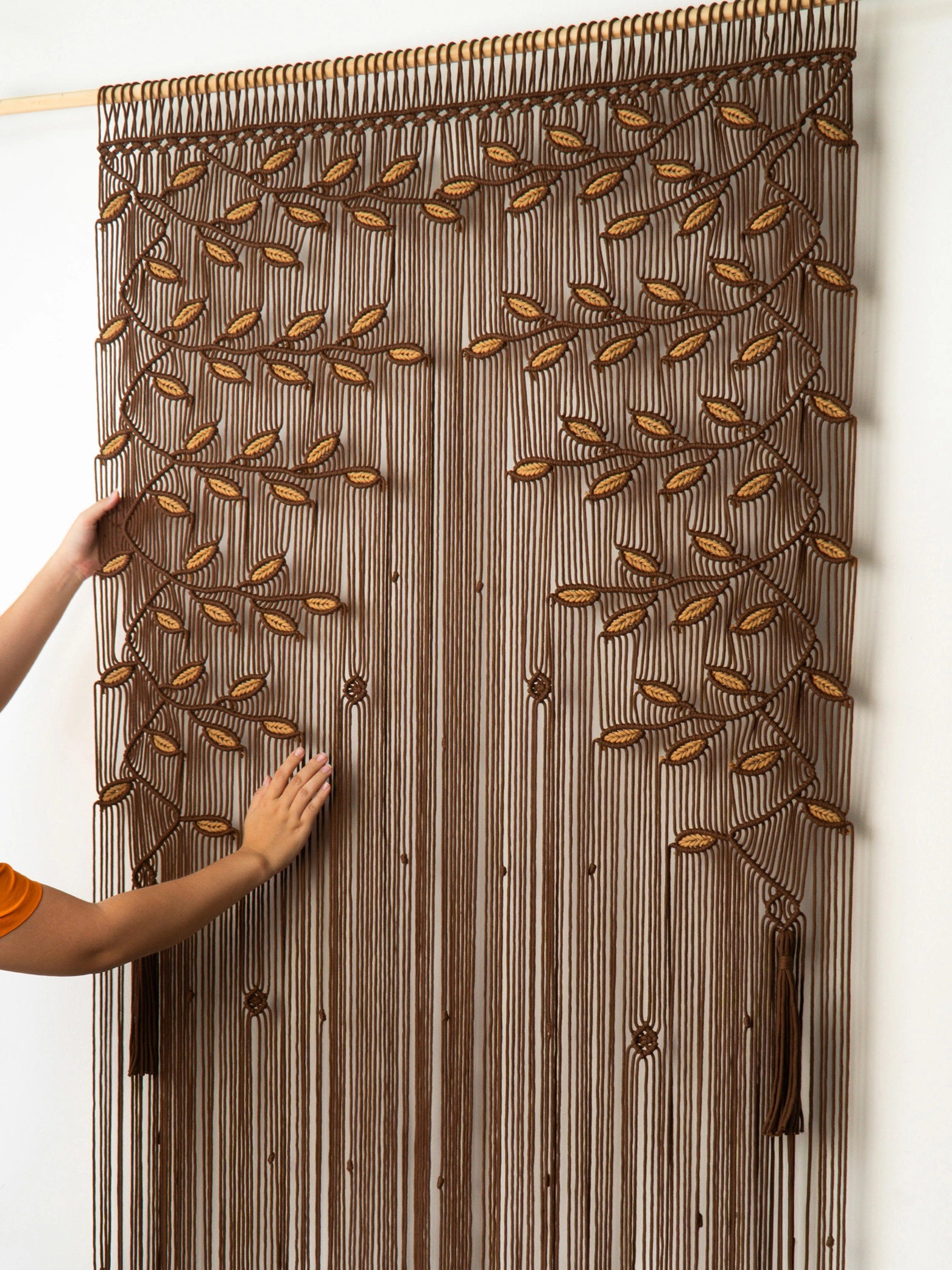 A brown and beige woven curtain panel with a tree branch and leaf pattern, shown hanging on a wall with a person's hands adjusting it on the left side.