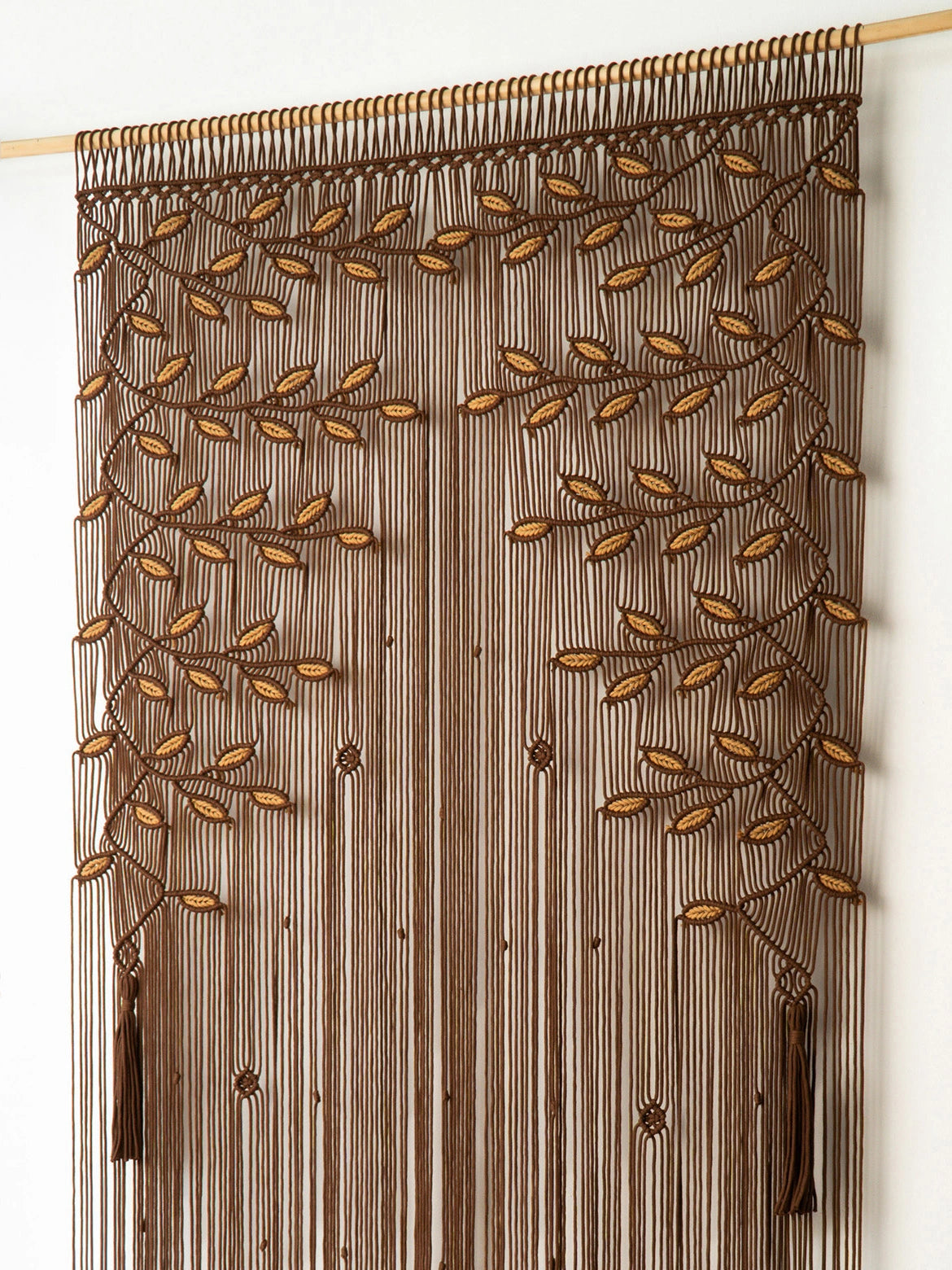 A brown beaded curtain featuring a leafy vine pattern woven with brown and tan beads, hanging from a wooden rod against a plain white wall.