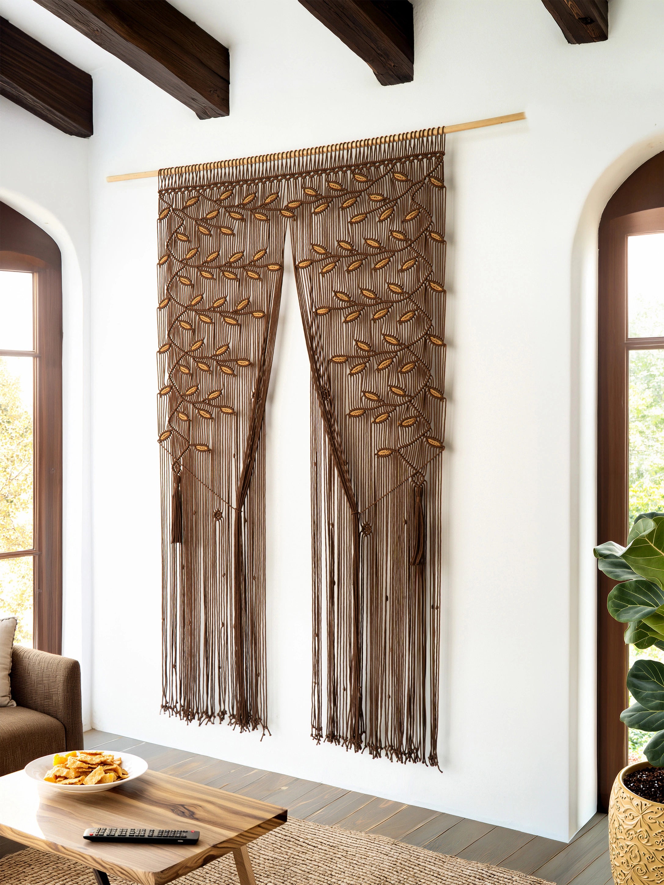A brown sheer curtain panel with embroidered leafy branches and long fringe, hanging on a wooden rod in a bright room with white walls, dark wood ceiling beams, and arched windows.