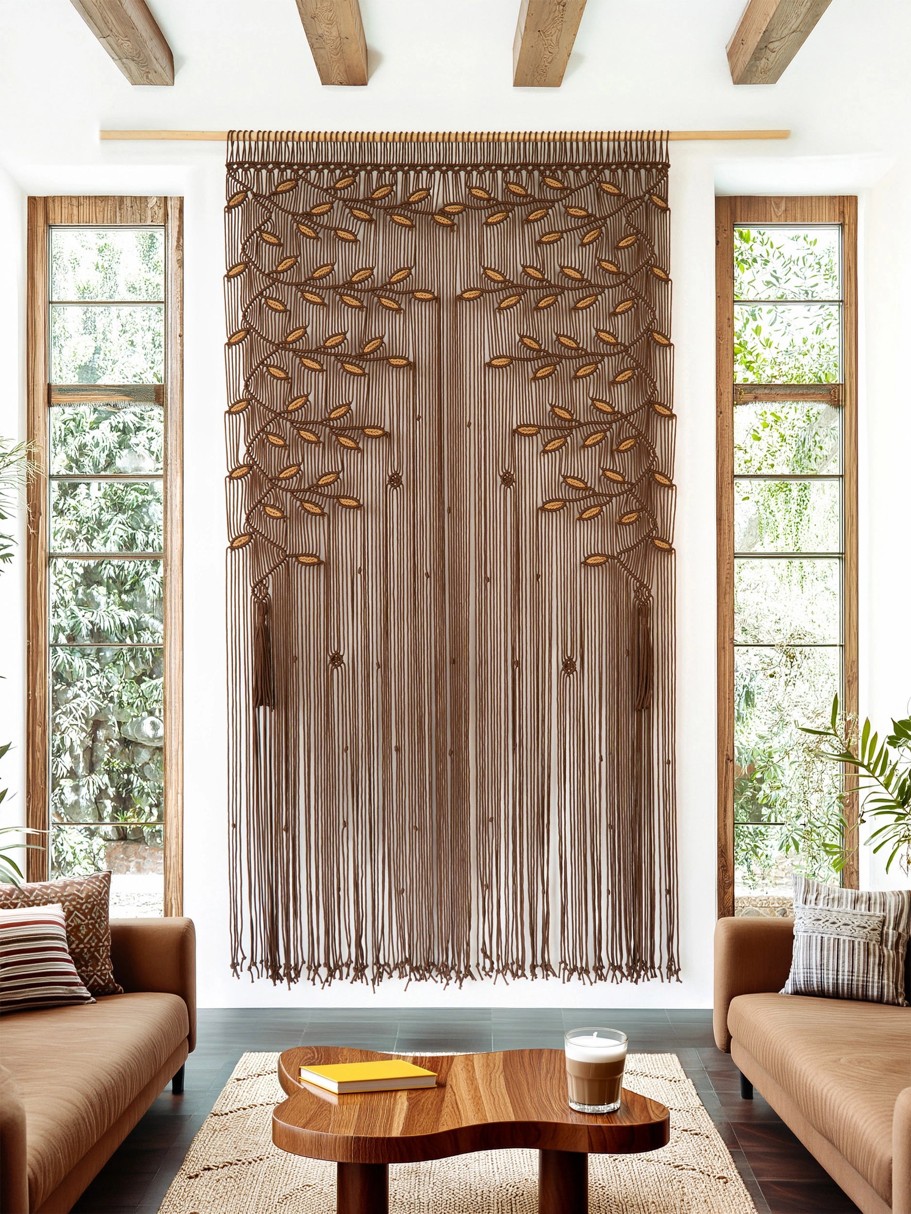 A brown macrame curtain panel with leafy vine patterns hangs in a window in a modern living room setting with brown leather sofas and a wooden coffee table.