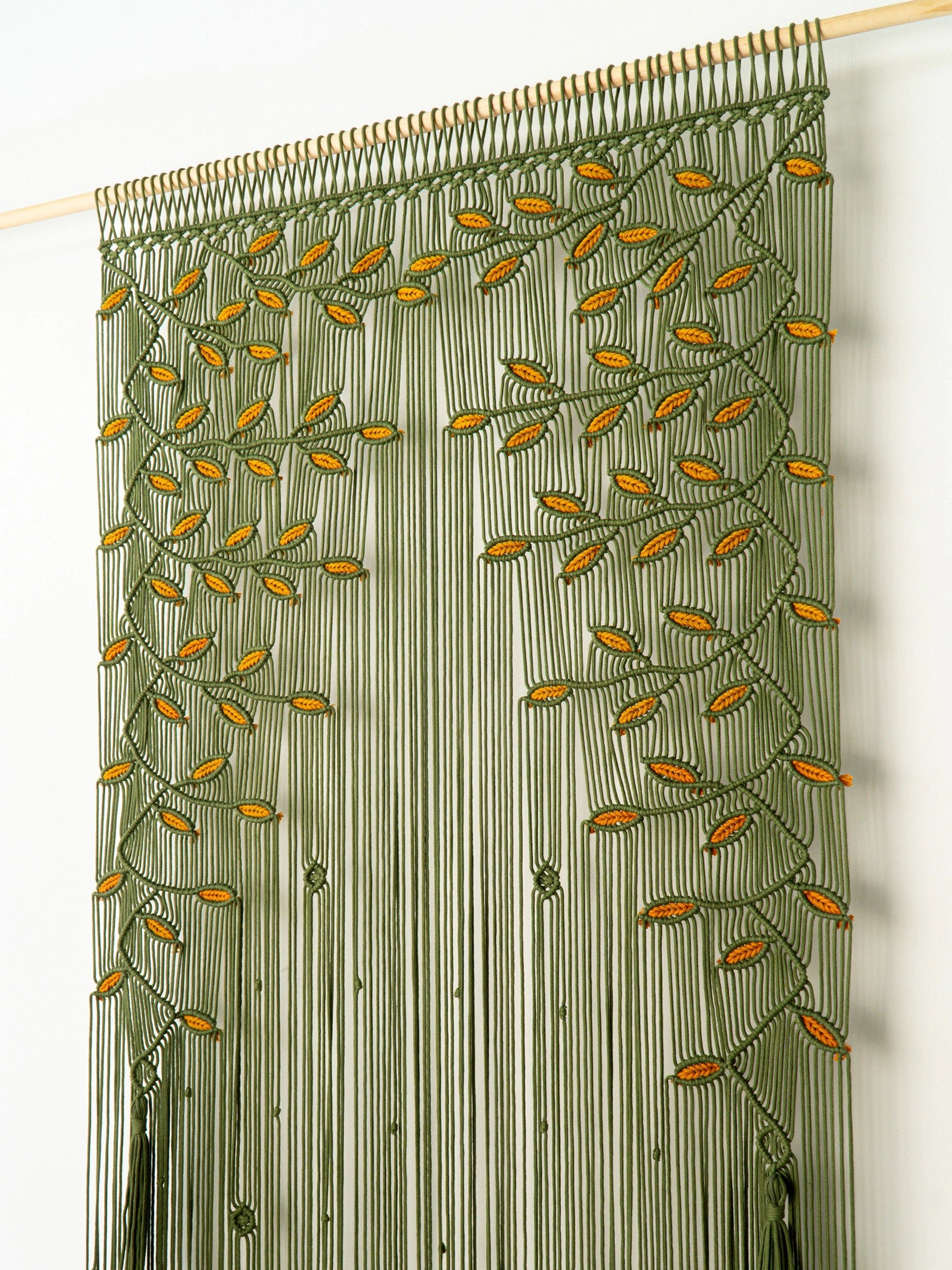 An olive green beaded curtain panel featuring a vine and leaf pattern with small orange-yellow accents, hanging from a wooden rod against a plain white wall.
