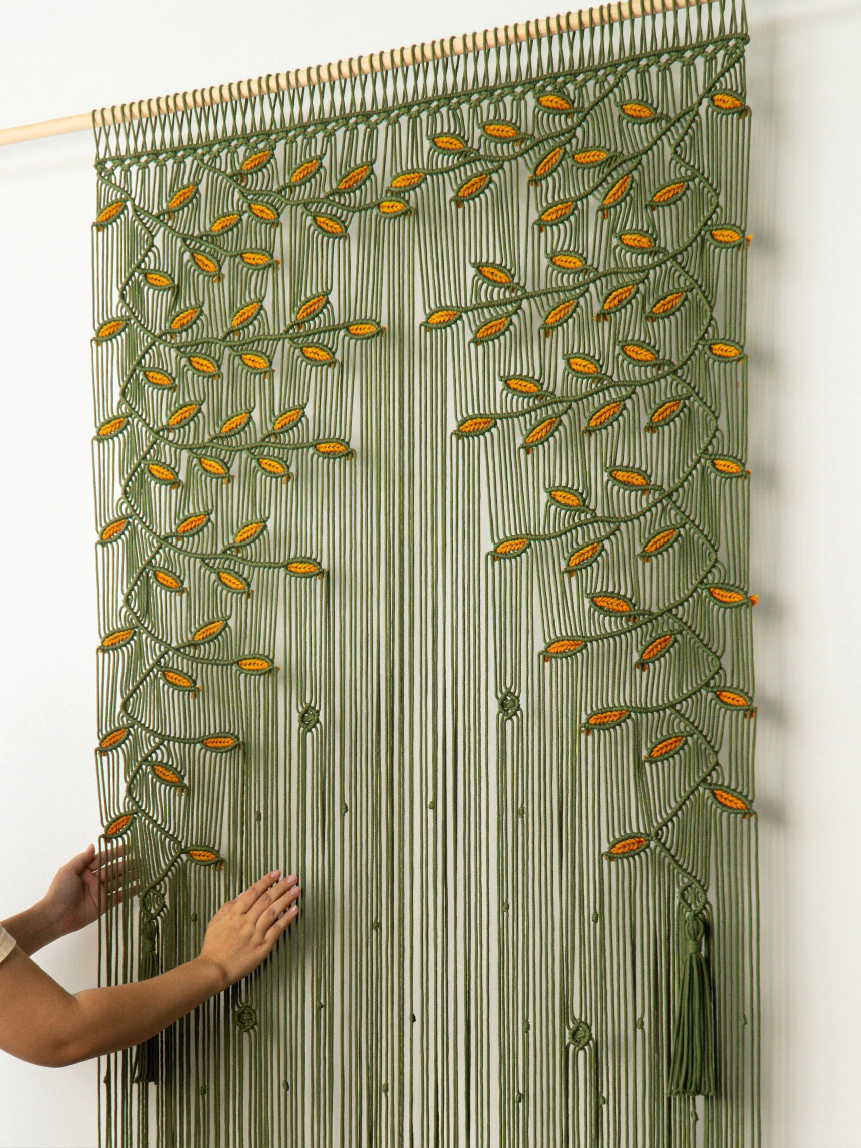 A green macrame curtain panel with a leafy vine pattern and tassels, hanging on a wooden rod against a white wall, with a person's hands adjusting it on the left.