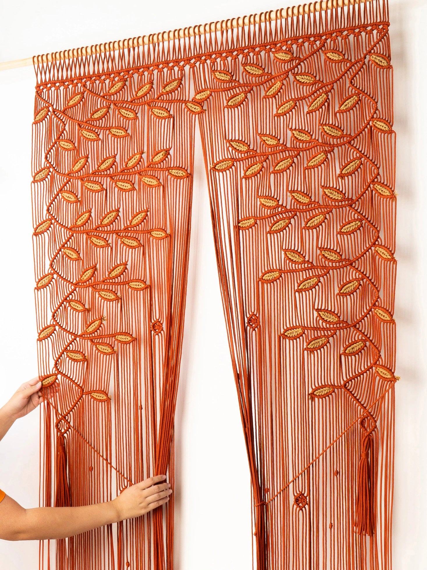 An orange and brown beaded curtain with leafy vine patterns, shown hanging on a rod with a person's hands holding it open against a white wall.