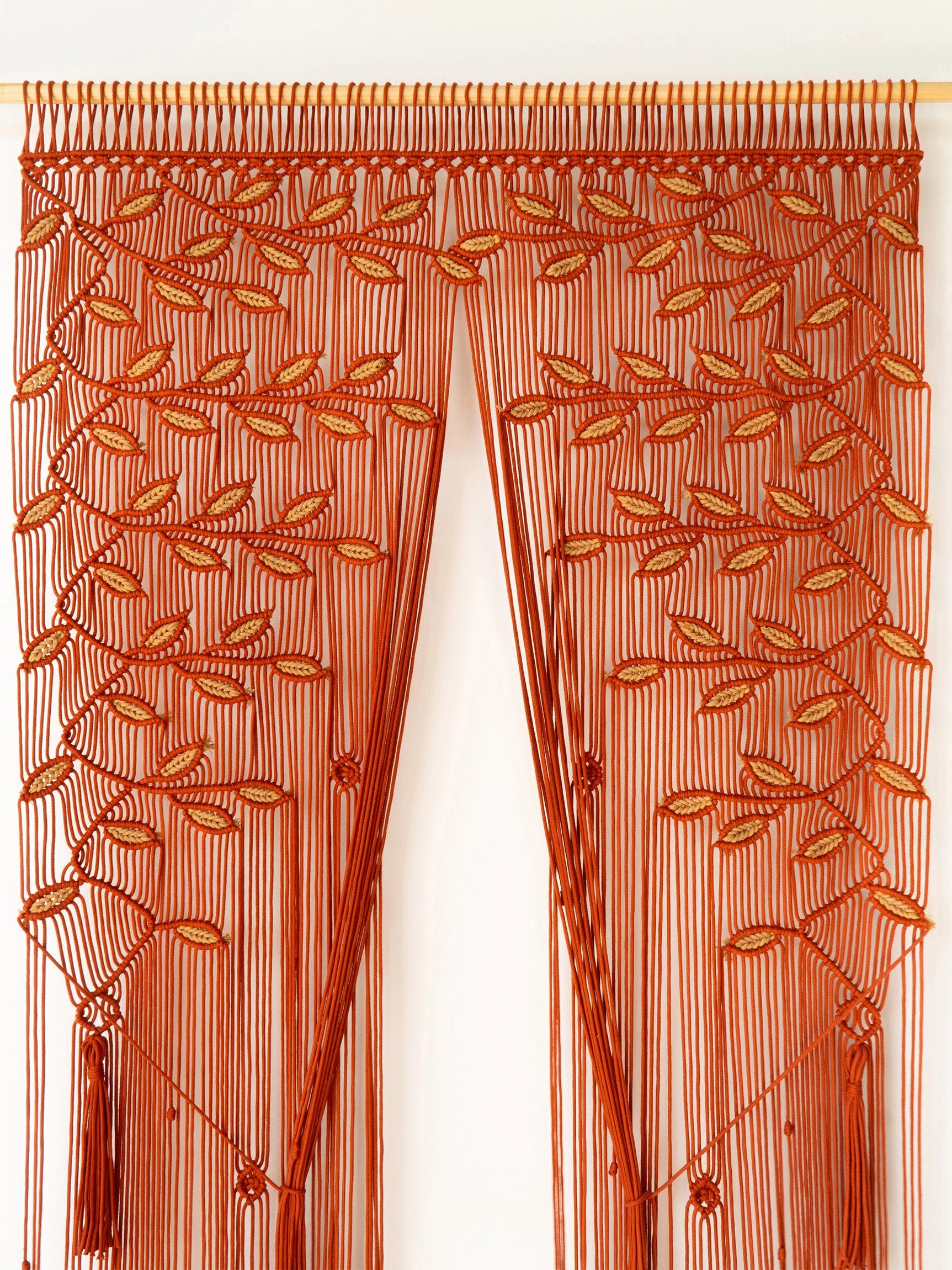 A rust-orange macrame curtain panel hanging from a wooden rod, featuring intricate woven leaf patterns and long fringe tassels against a plain white background.