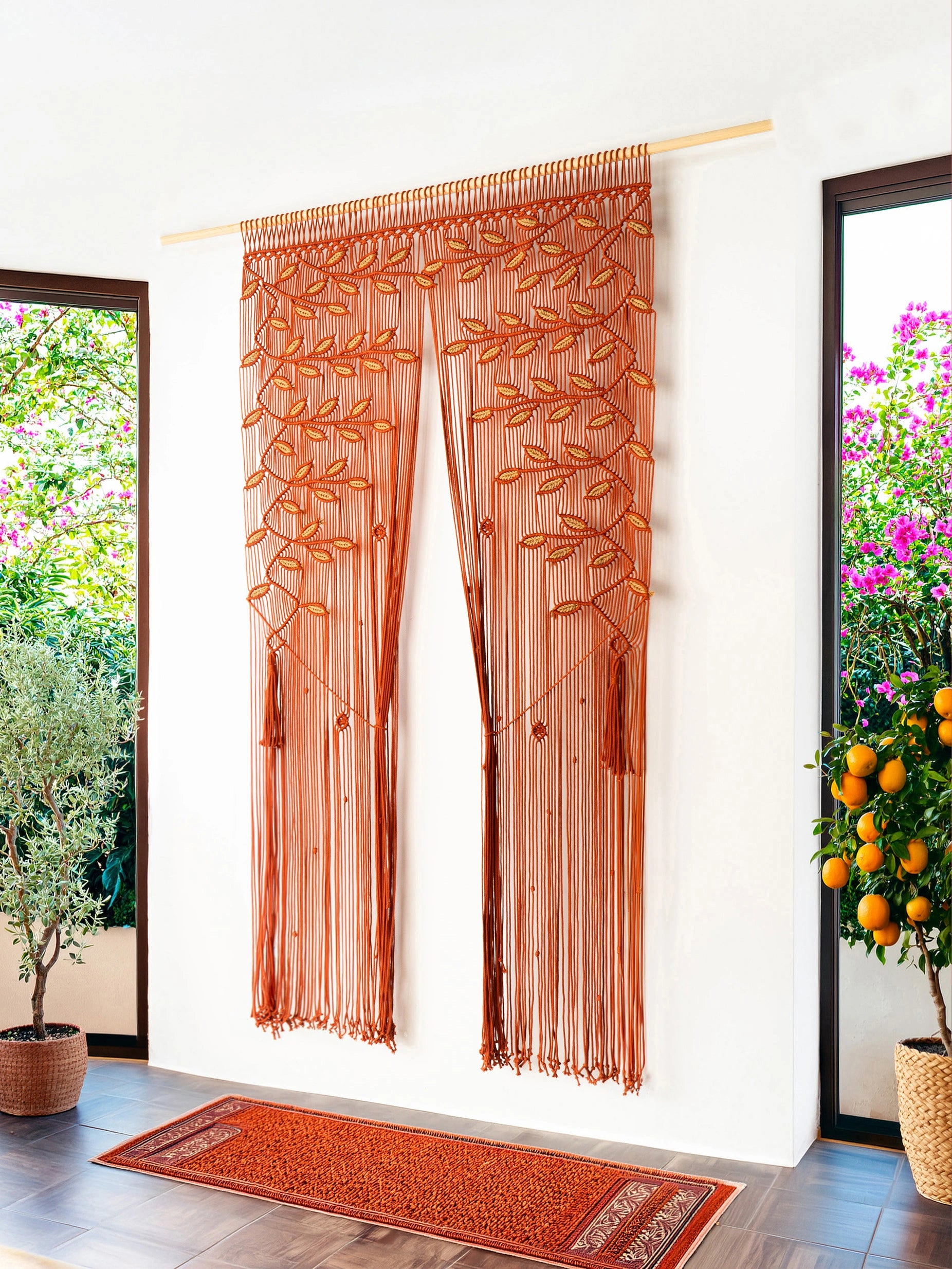 A rust-orange sheer curtain panel with embroidered leaf and vine patterns, hanging on a light wood rod in a bright room with potted plants and a matching runner rug on the floor.