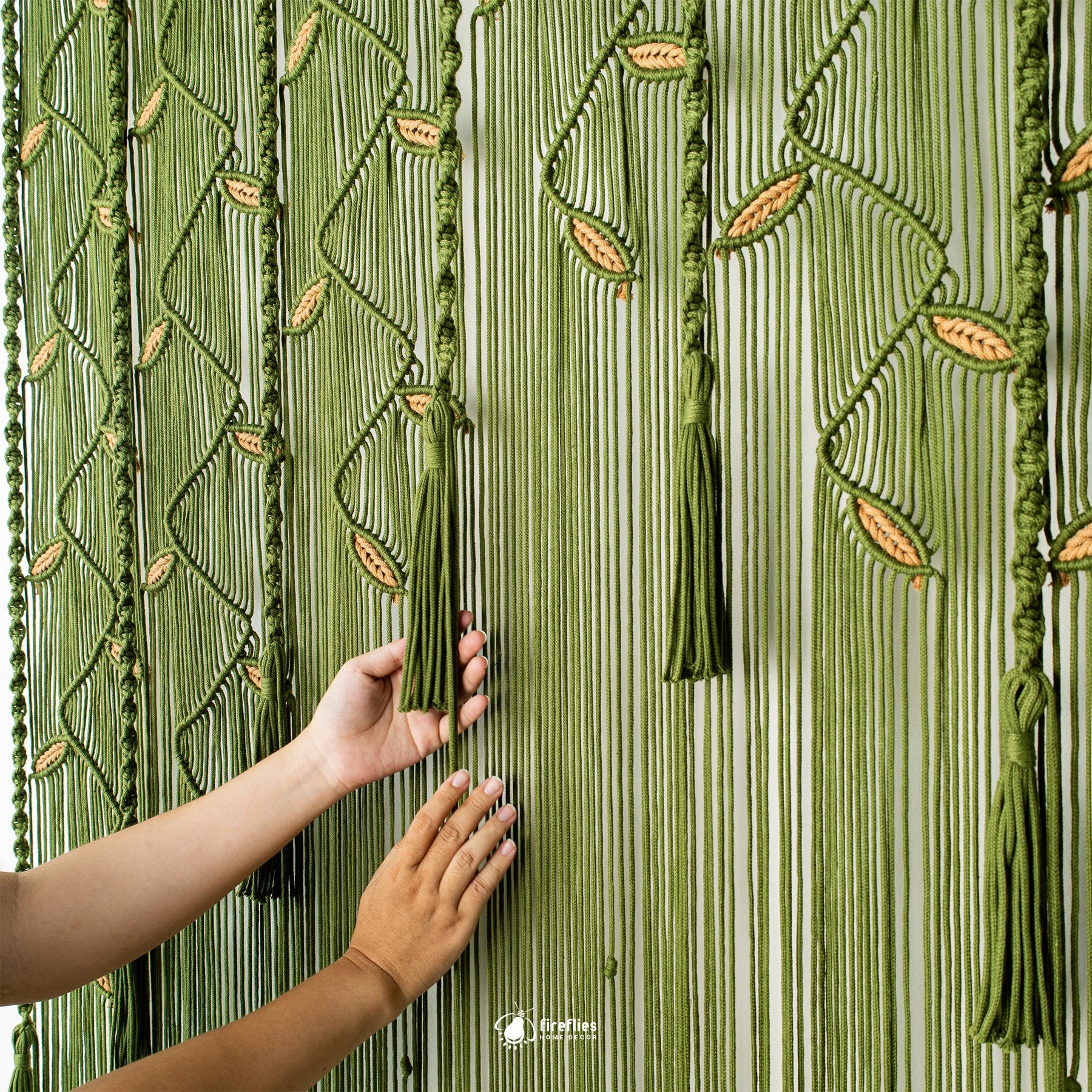 A close-up of a green macrame curtain with vertical woven stripes, hanging tassels, and leaf patterns, with two hands holding the curtain open. A small 'The Art of Living' logo is visible at the bottom center.