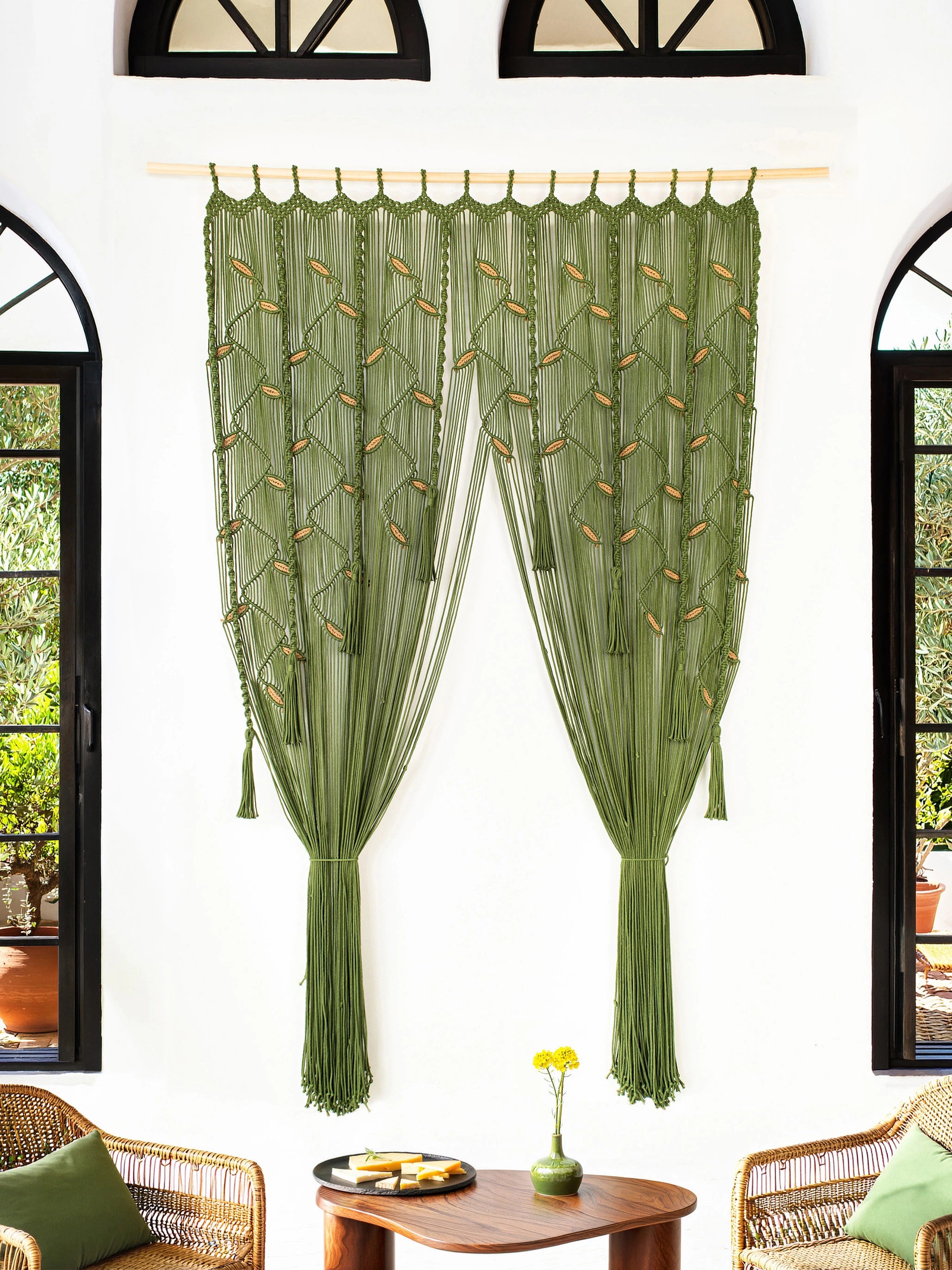 Olive green sheer curtain panels with embroidered leaf patterns, hanging on a wooden rod in a bright room with arched windows and wicker furniture.