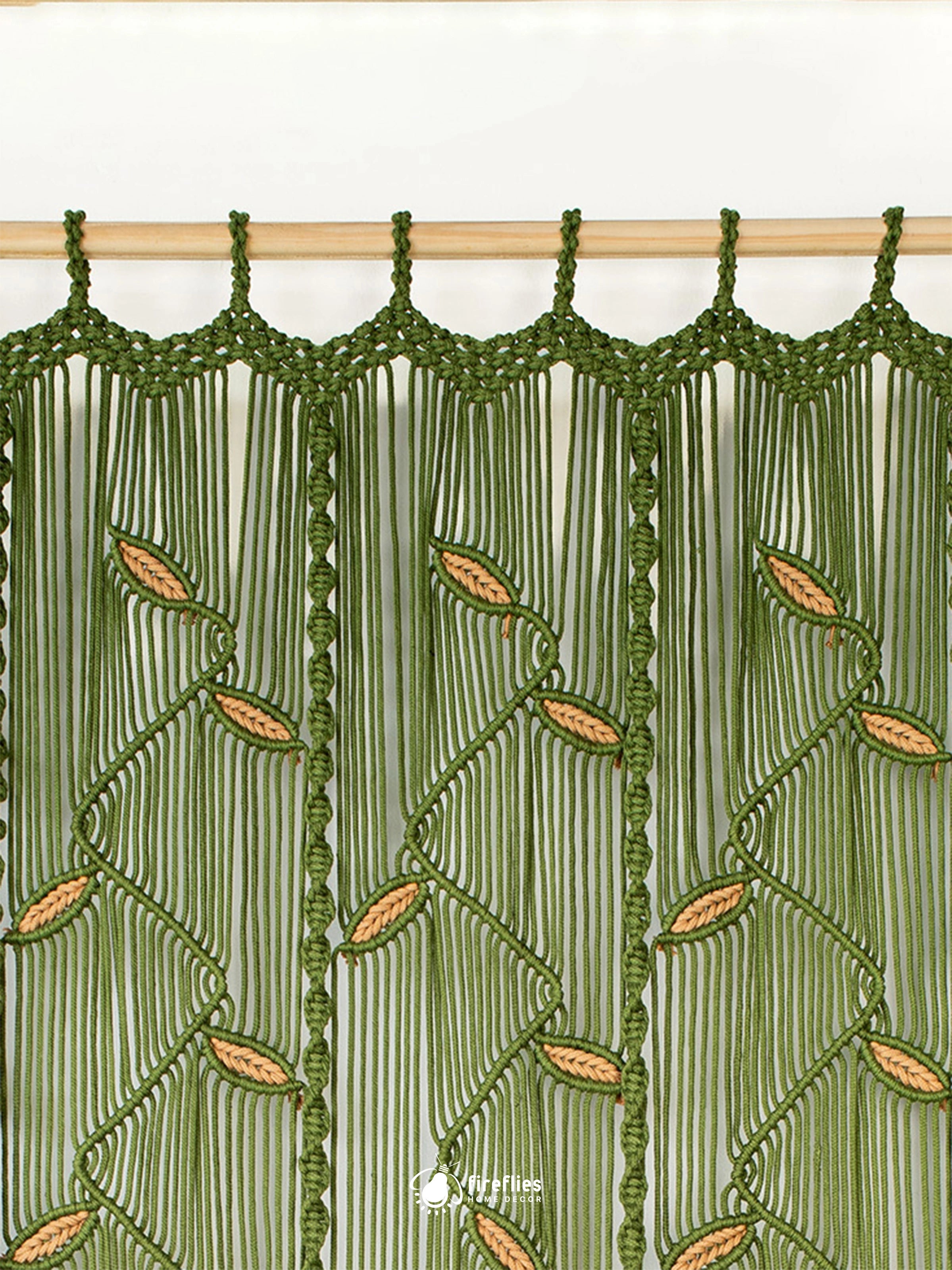 A close-up of a green macrame curtain panel hanging from a light wood rod. The design features vertical strands of green cord with knotted leaf patterns in a warm tan/beige color.