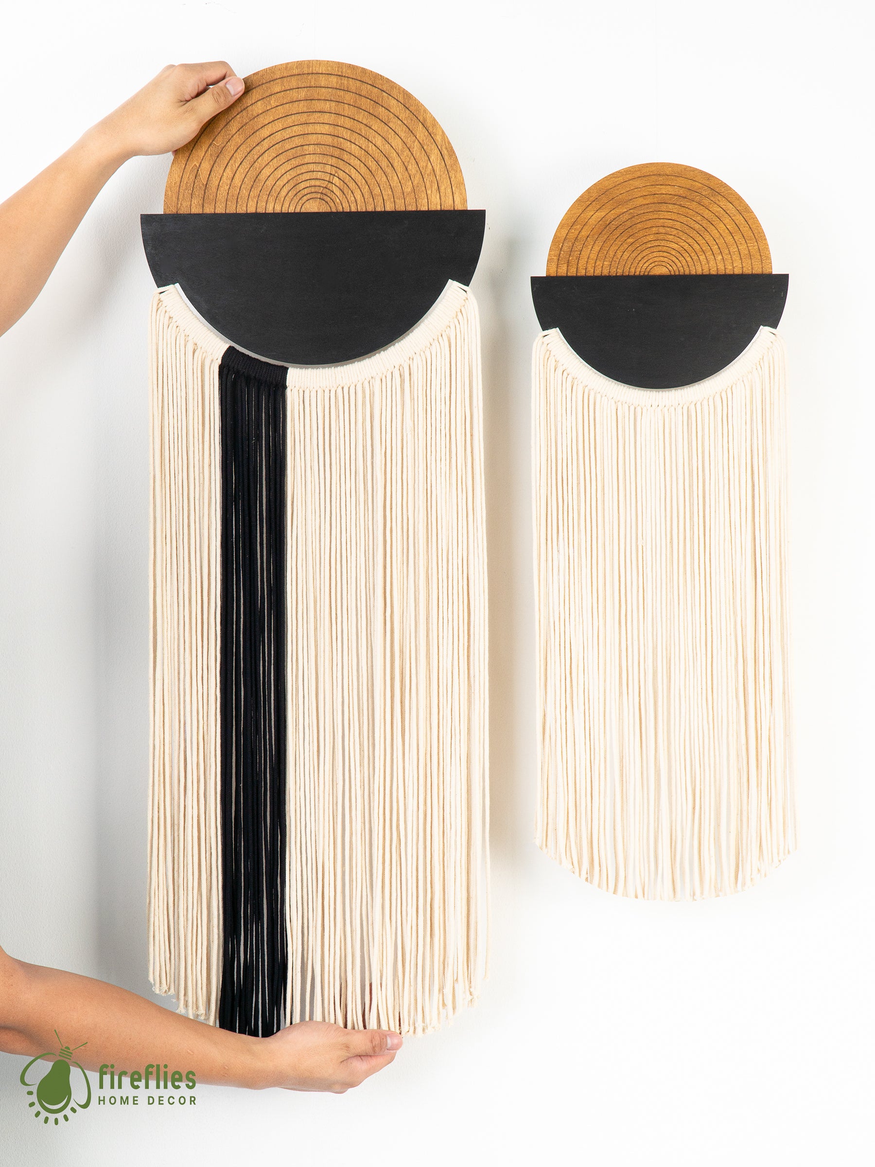 Two wall hangings with woven designs and black accents, one held by a hand, on a white background.