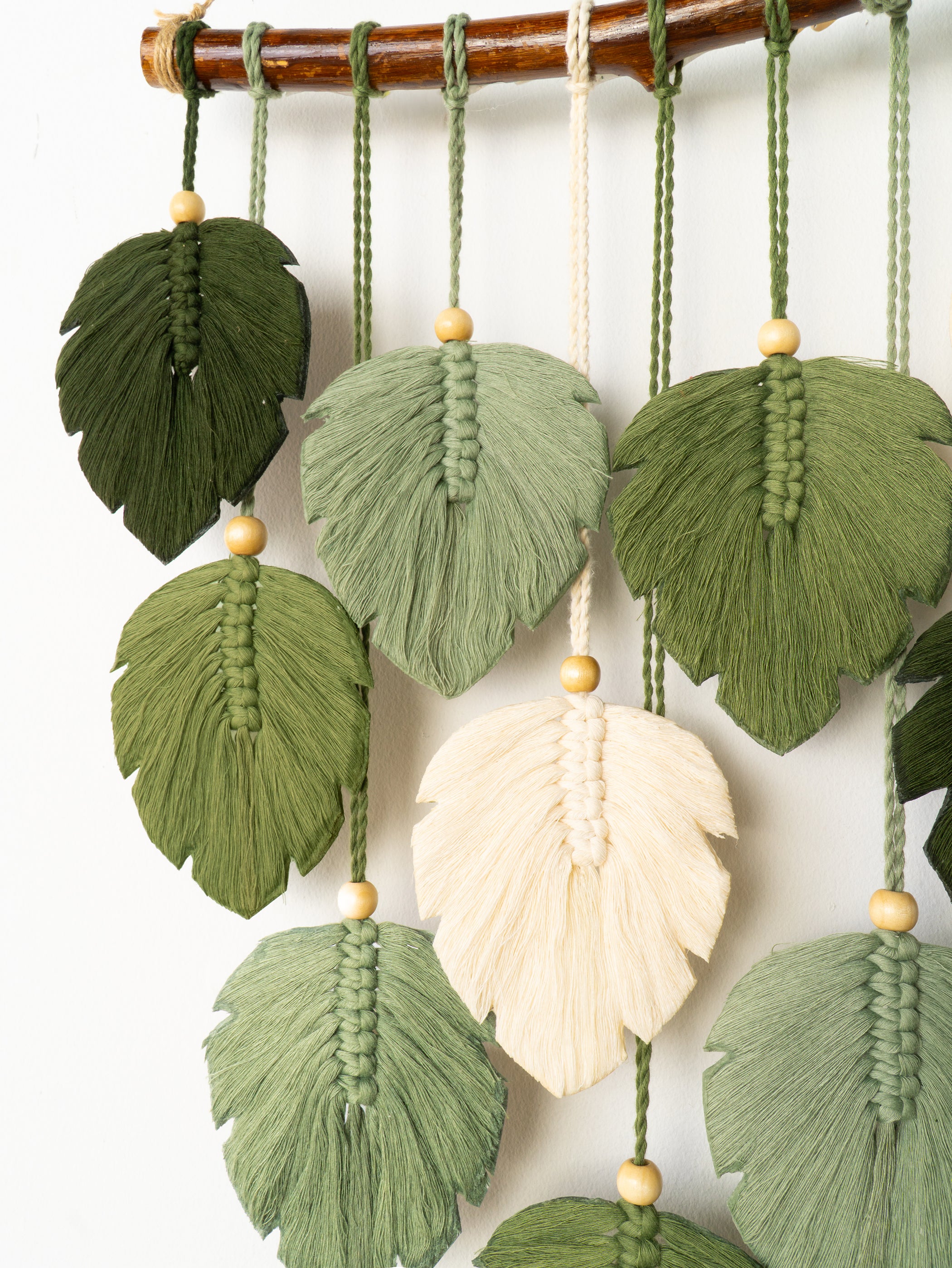 Decorative wall hanging with green leaf-shaped tassels and a central cream-colored feather on a white background.
