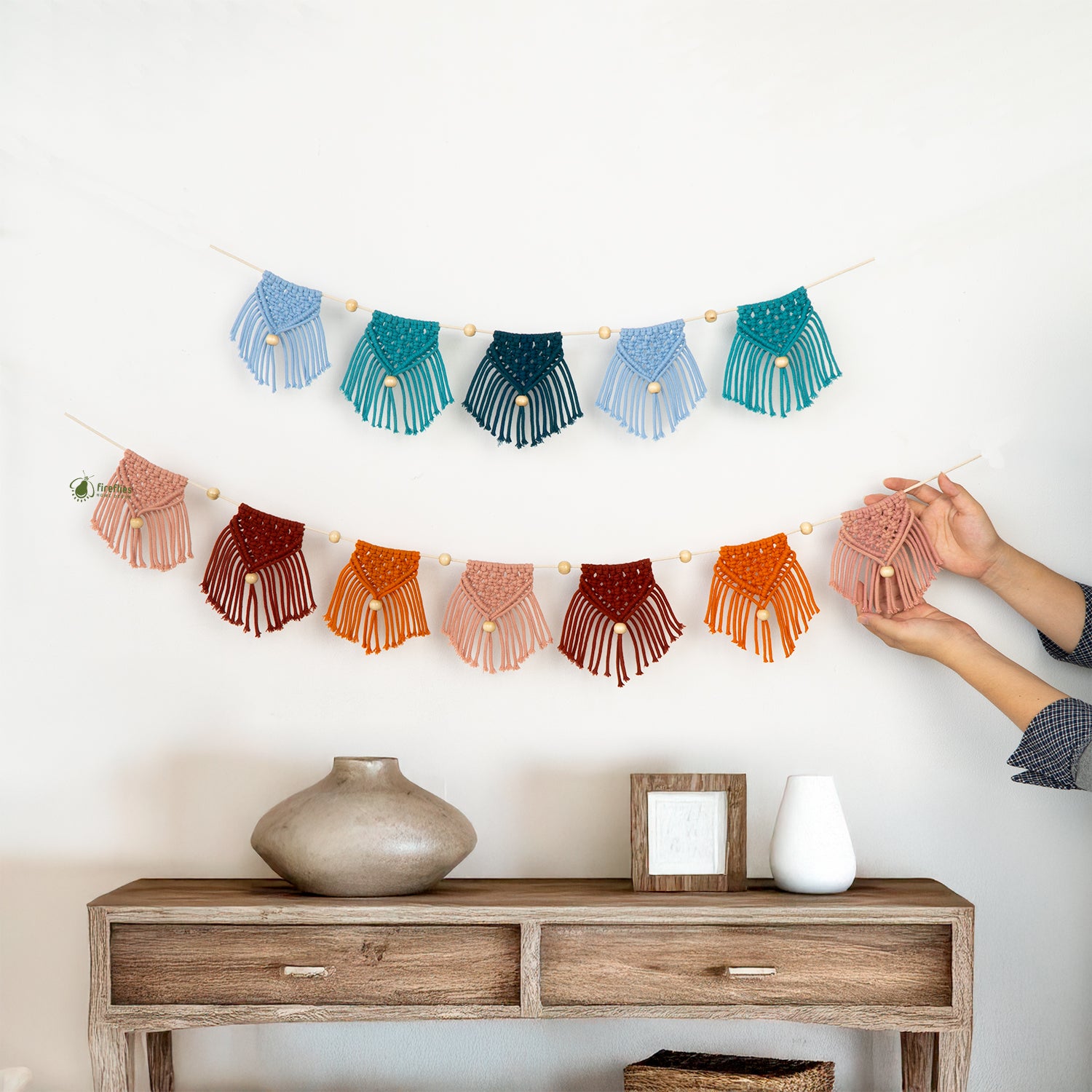 Colorful macrame wall hanging being adjusted above a wooden console table with decorative items.