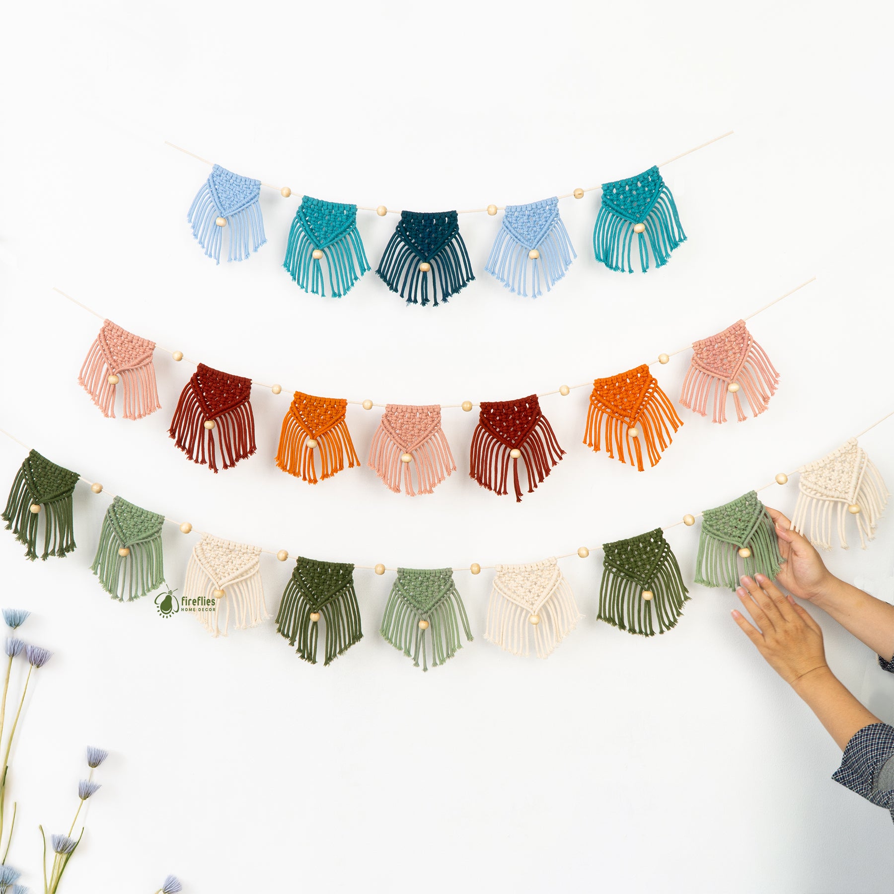 Colorful macrame garland with a hand reaching to touch one of the hanging pieces on a white background.