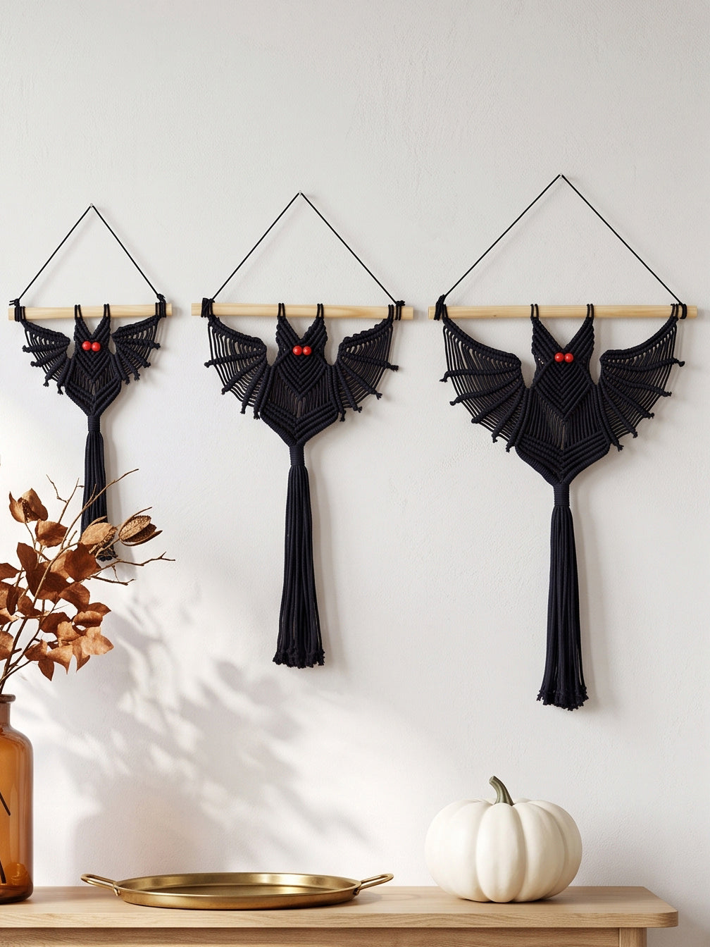 Set of three jet black macrame bat wall hangings in graduated sizes displayed on a white wall above a wooden entryway console table.   