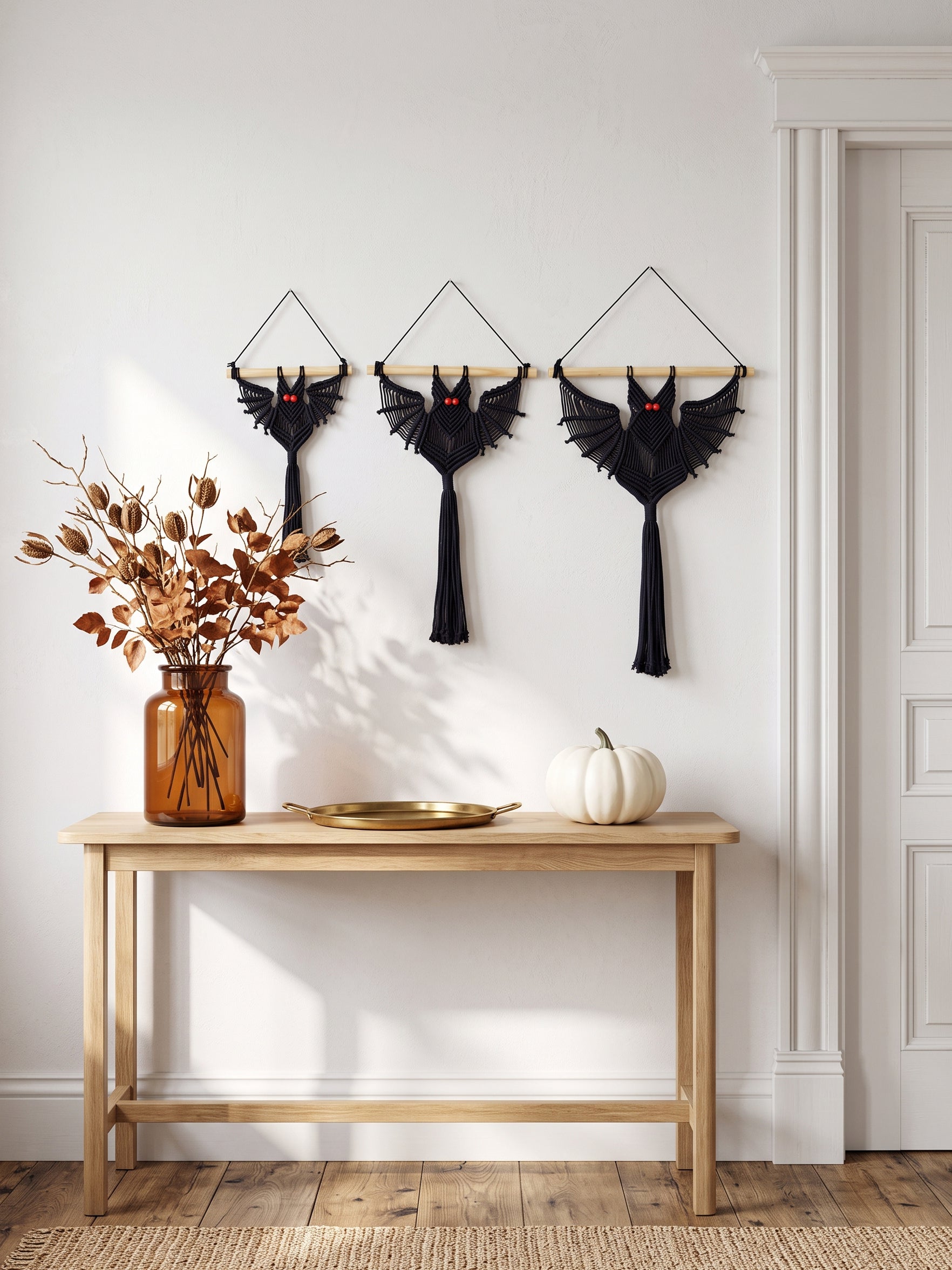 Trio of spooky macrame bats hanging in a row above a brass tray, dried autumn botanicals, and a whit pumpkin.   
