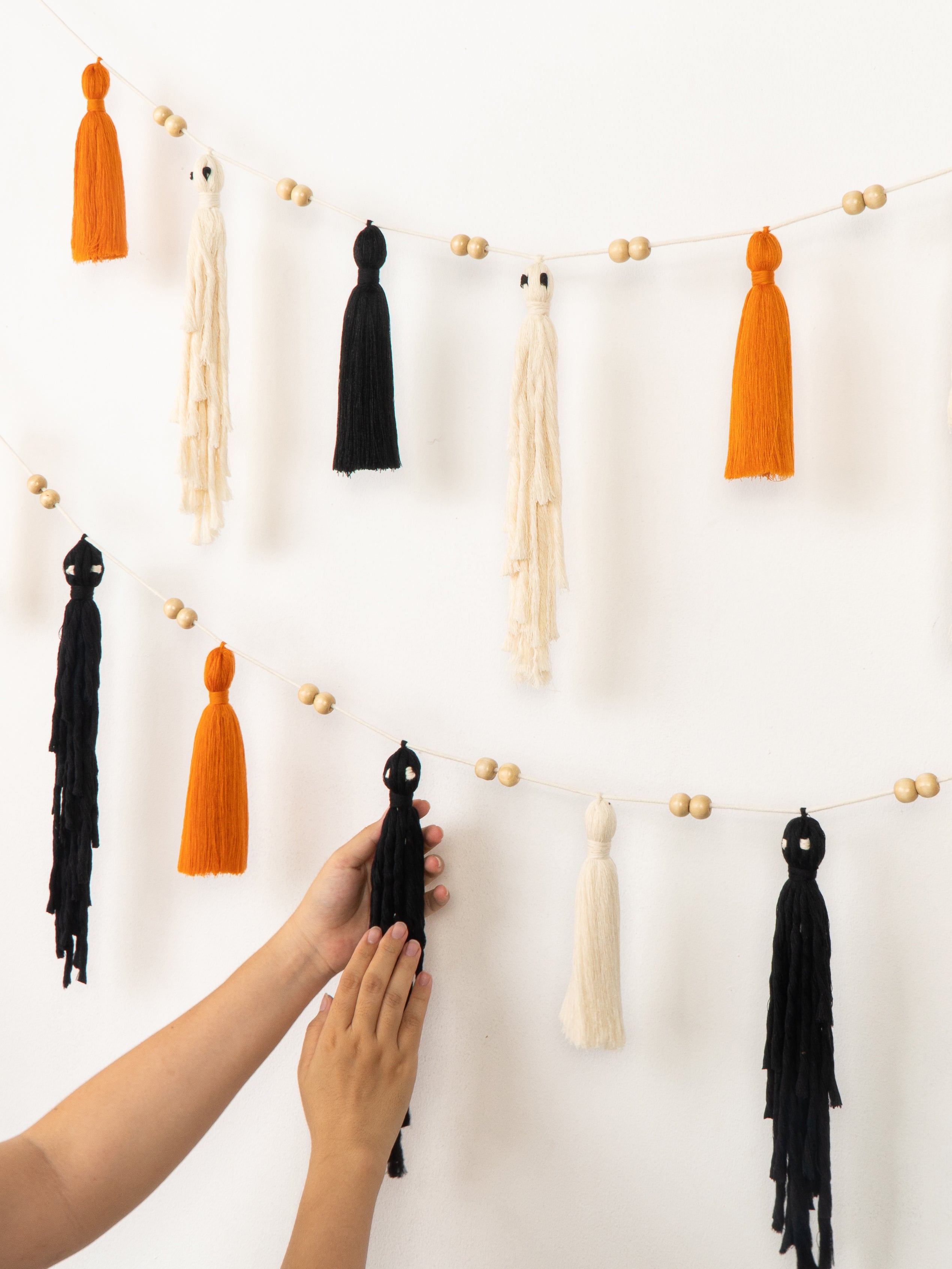 A string of decorative tassels and small wooden beads hanging on a white wall, with a person's hands adjusting one of the black tassels.