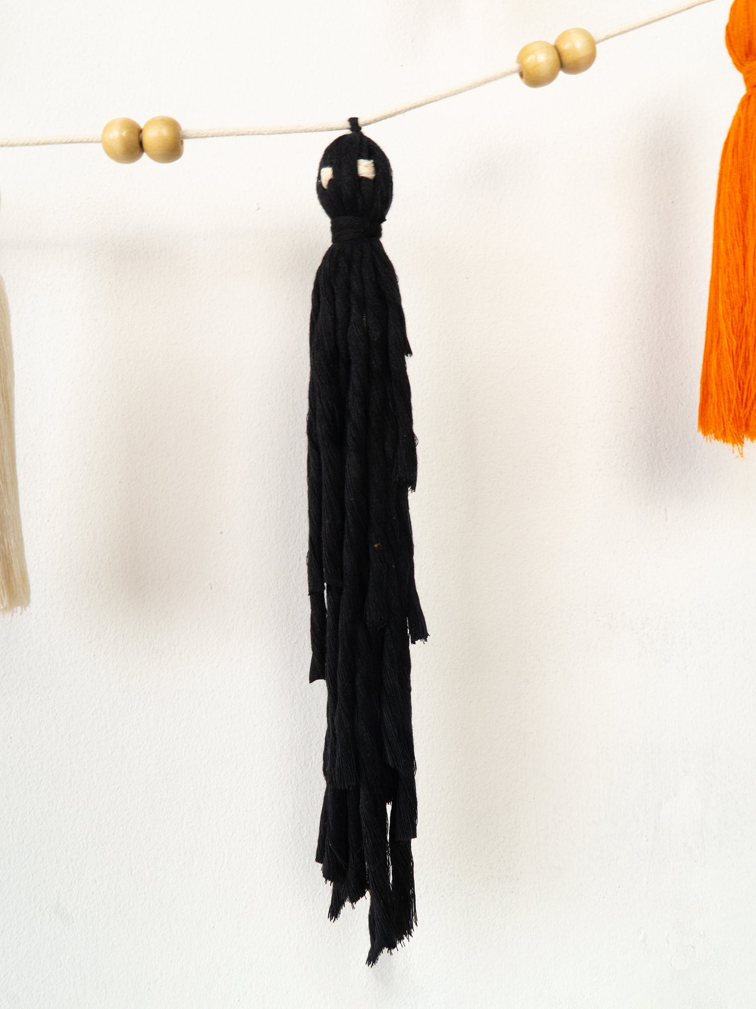 A black tassel garland hanging from a string with wooden beads, shown against a plain white wall with partial orange and cream tassels visible at the edges.