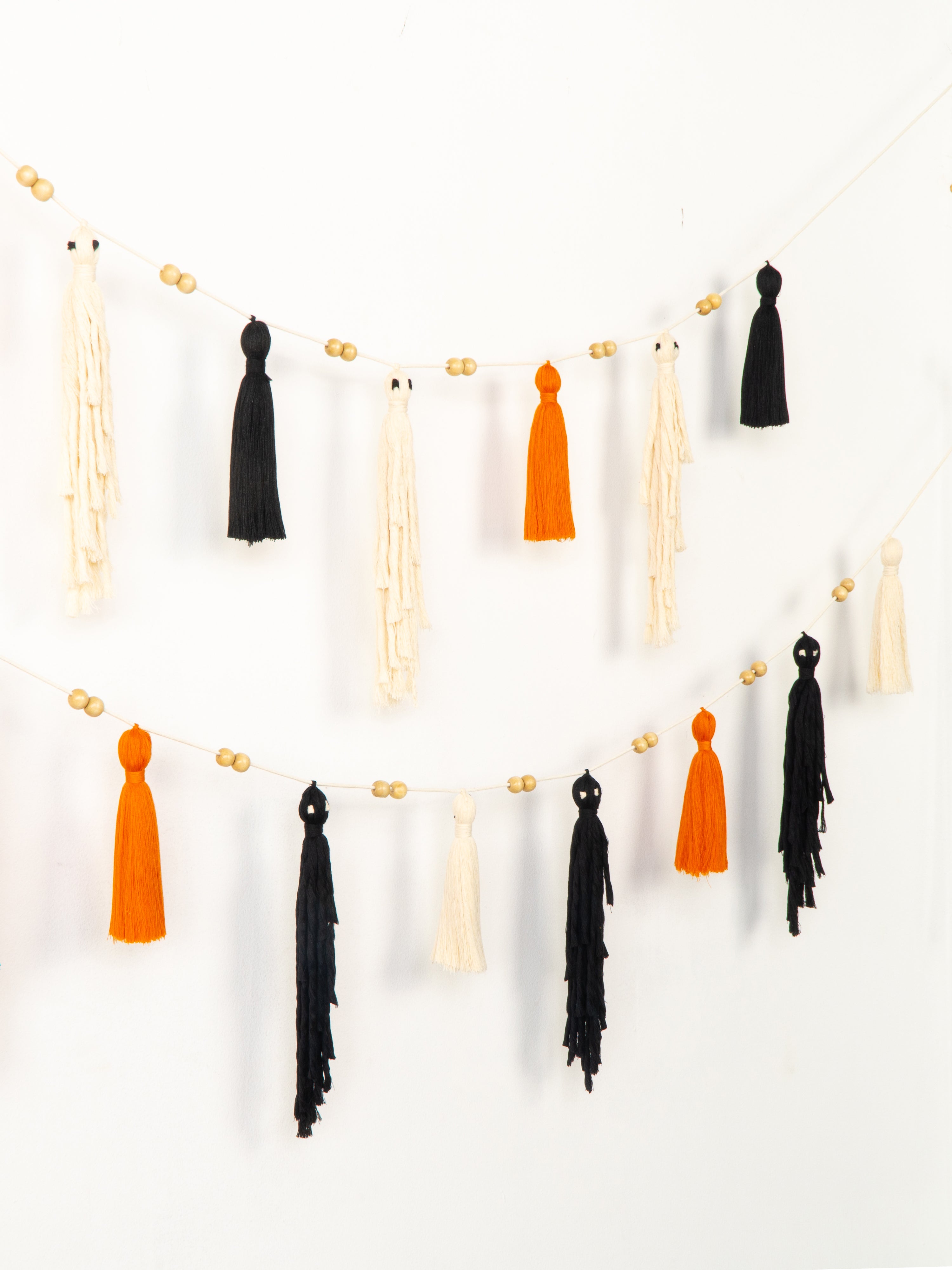 A boho-style tassel garland hanging in two curved rows against a plain white wall. The garland features black and orange tassels alternating with cream tassels, accented by small round wooden beads on a white string.