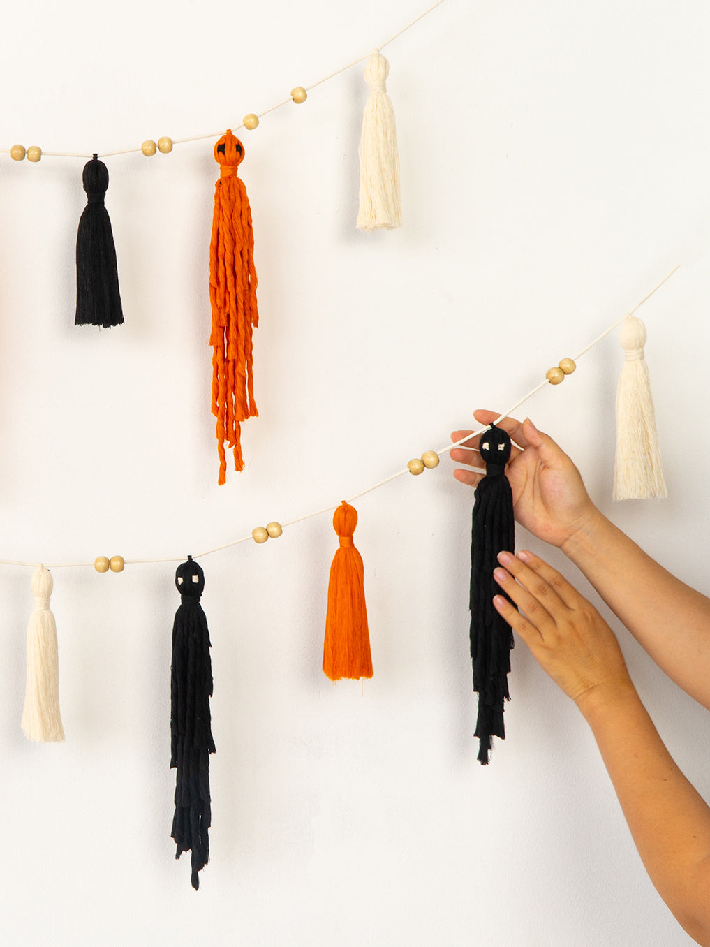 A string garland of knotted tassels in black and orange with small gold beads, shown hanging on a white wall with a person's hands adjusting one of the black tassels.