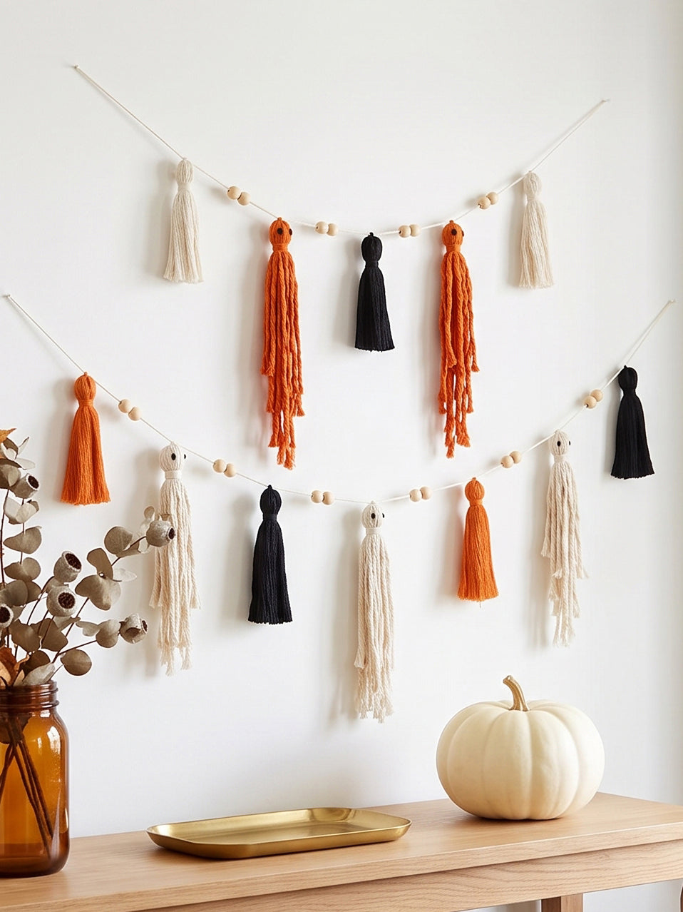 Pumpkin-themed tassel garland with orange, black, and cream tassels hanging on a string against a white wall, styled above a wooden console table with a white pumpkin, gold tray, and eucalyptus in an amber vase.