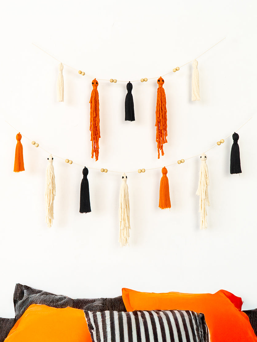 A bohemian-style tassel garland with orange, black, and cream tassels hanging in two rows on a white wall above a bed with matching orange and striped pillows.