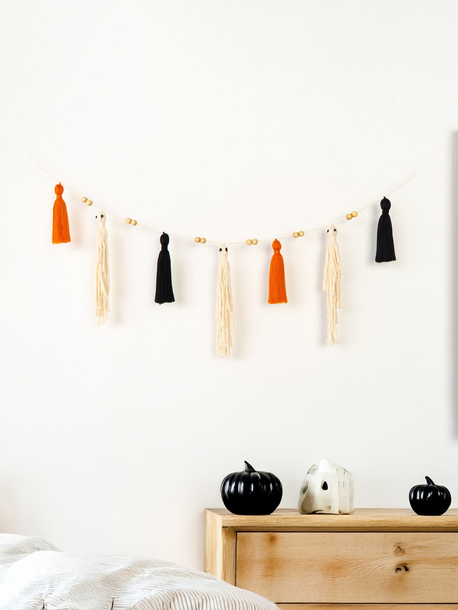 A Halloween-themed tassel garland with orange, black, and cream tassels hanging on a string against a white wall, with a light wood dresser below holding small black and white pumpkin decorations.