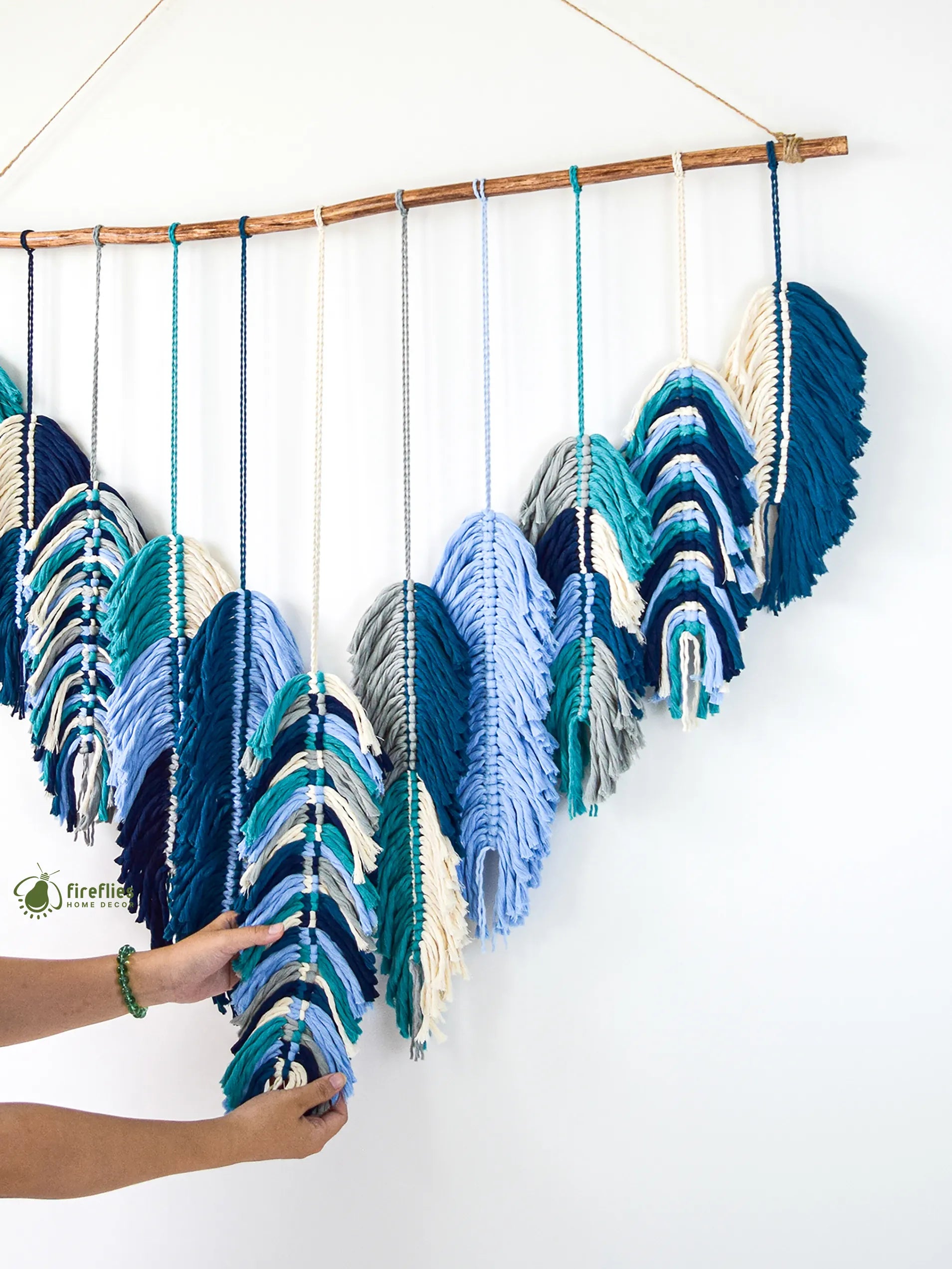 Decorative wall hanging with blue and white yarn on a wooden stick against a white background.