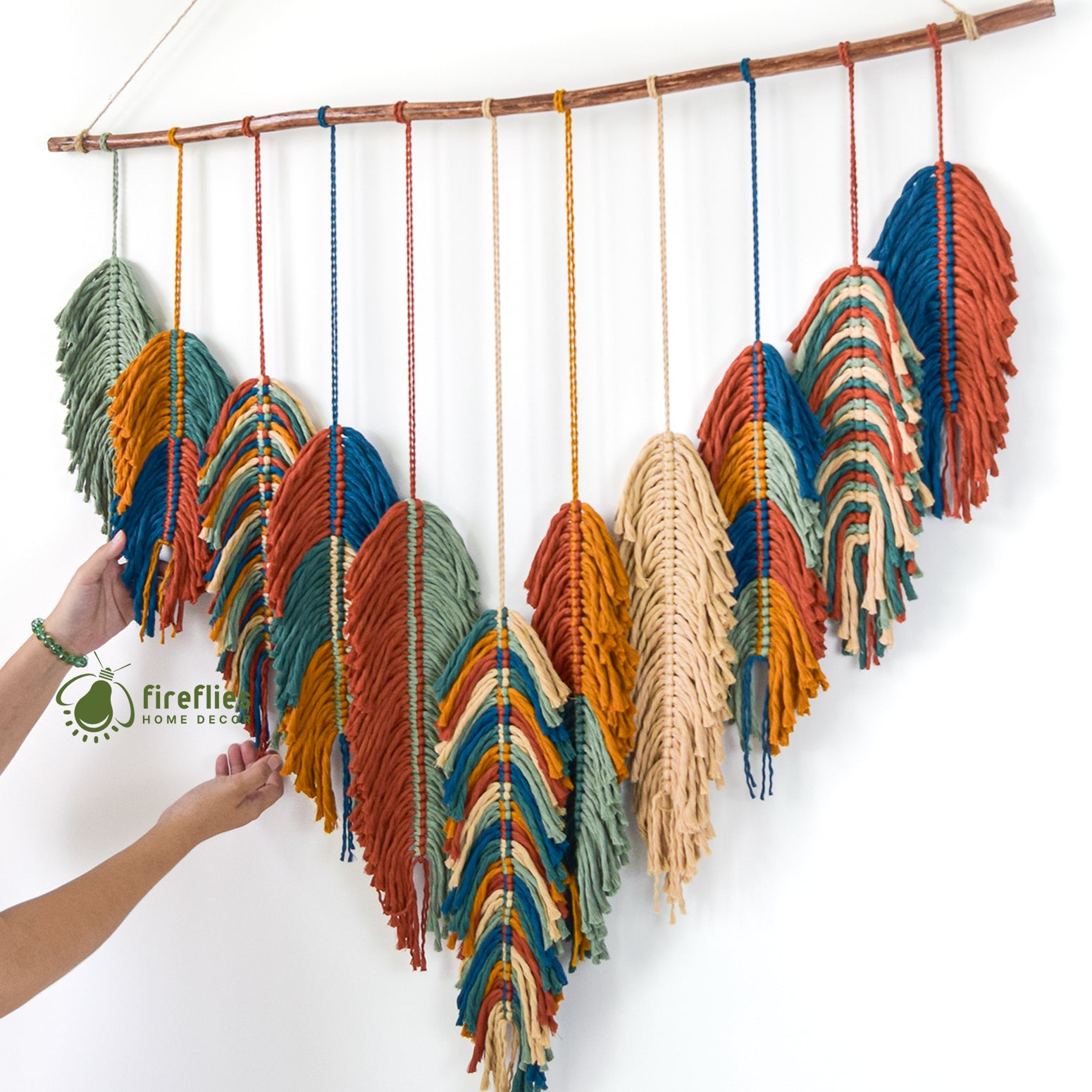 Colorful macrame wall hanging with a person's hands holding it against a white background, featuring the brand 'Firefly Home Decor'.