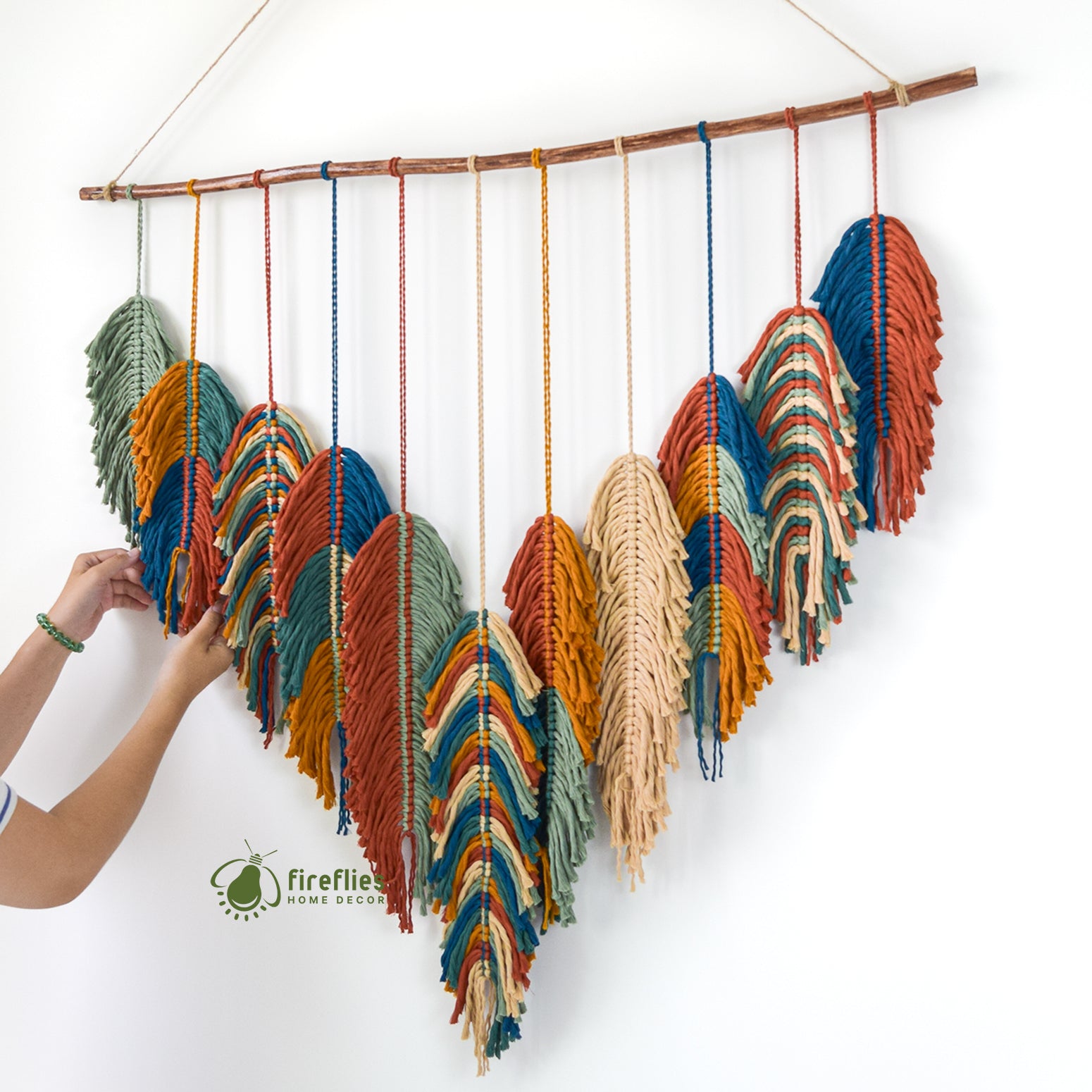 Colorful woven wall art with a person adjusting it against a white background, featuring the brand 'Fireflies Home Design'.