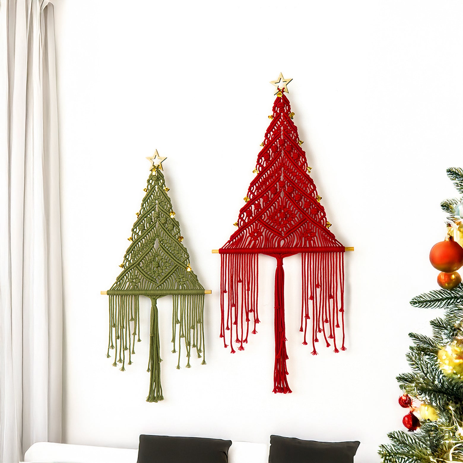 Two macrame Christmas trees, one green and one red, hanging on a white wall with a decorated Christmas tree in the corner.
