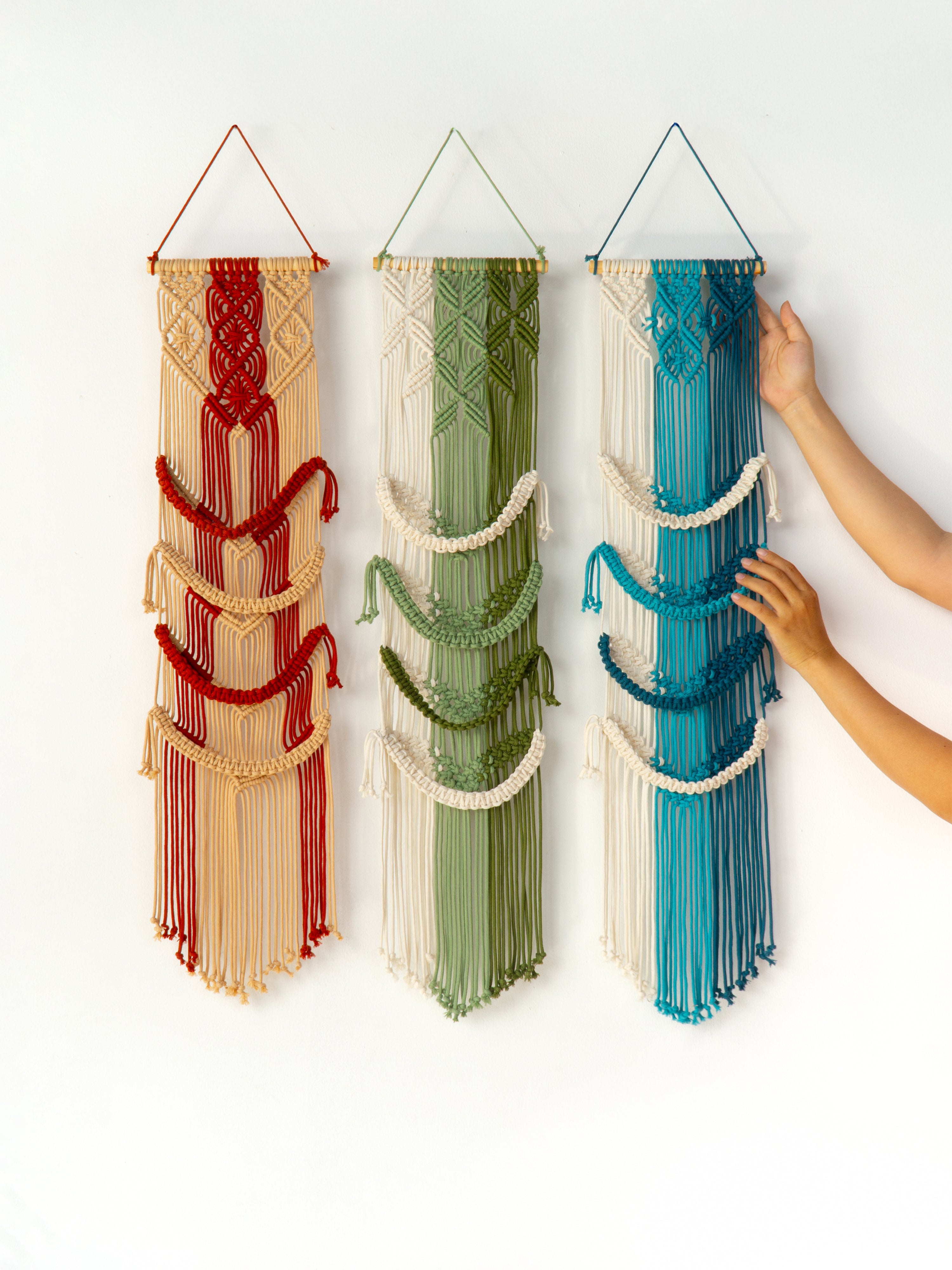 Three colorful wall hangings made of woven strips, with a hand reaching to adjust one of them.
