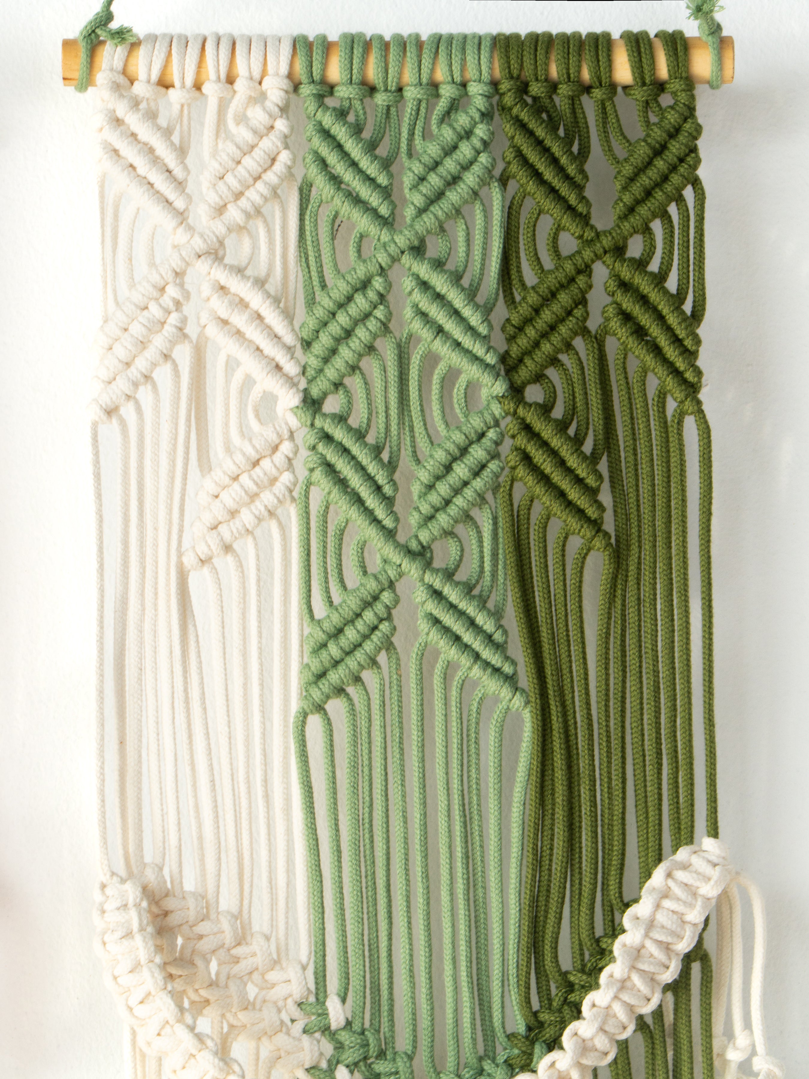 Macrame wall hanging with geometric patterns in white, green, and brown on a white background