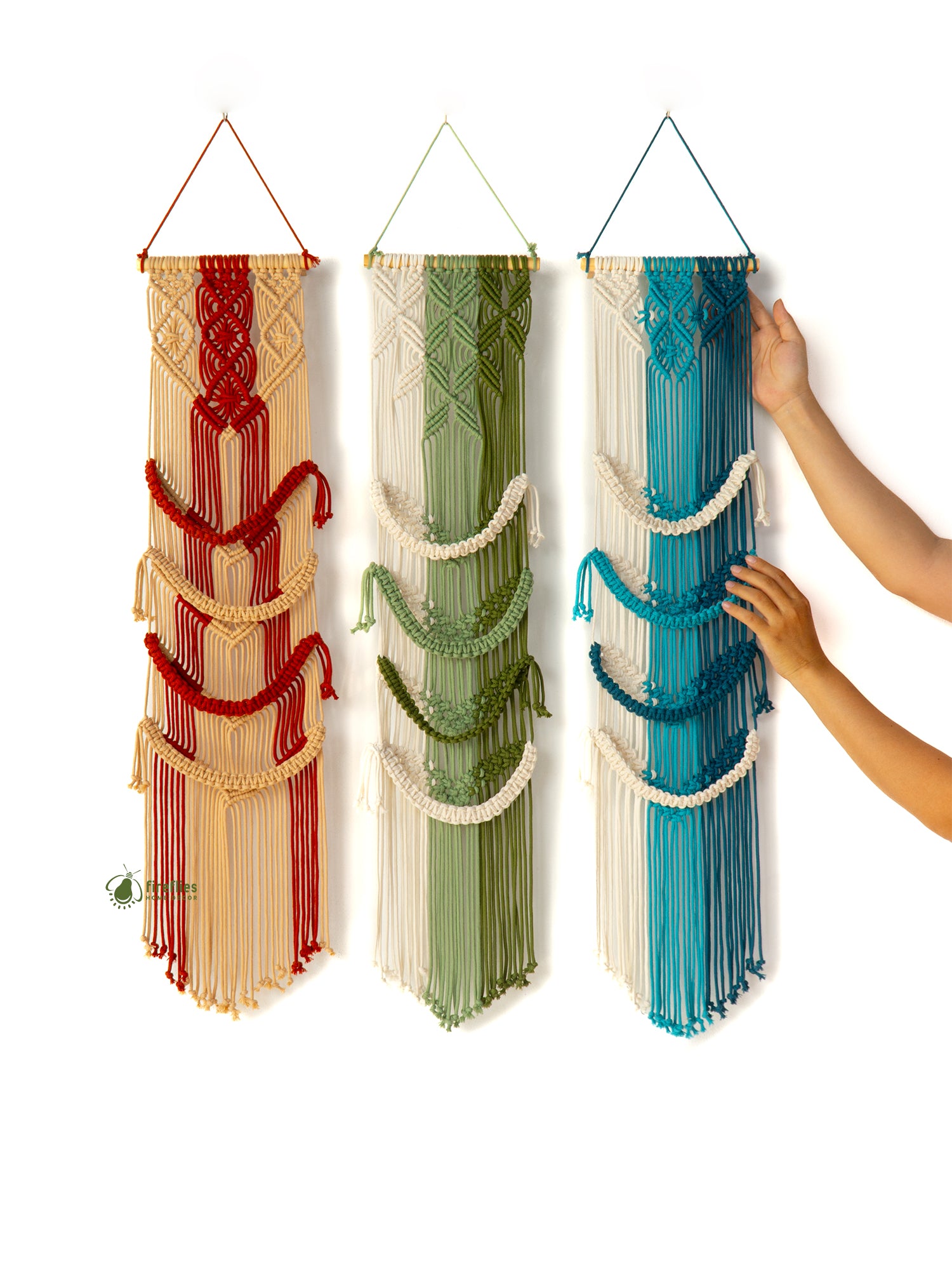 Three macrame wall hangings in red, green, and blue hanging on a white wall.