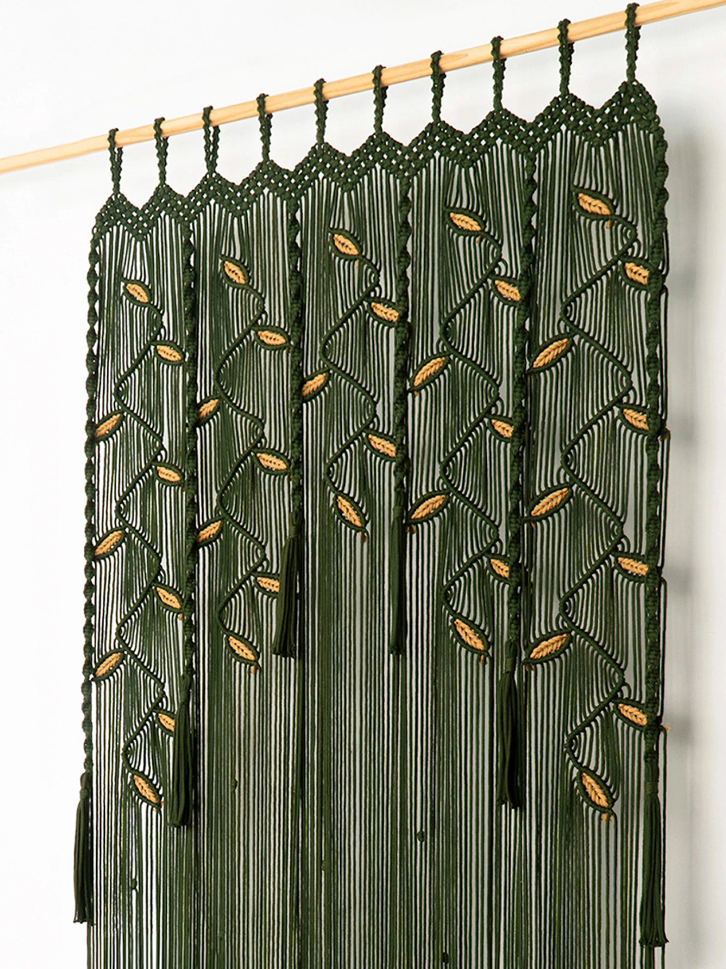 A dark green macrame curtain panel hanging from a light wood rod against a plain white wall. The panel features intricate woven patterns with small gold-colored leaf accents and long hanging fringe tassels.