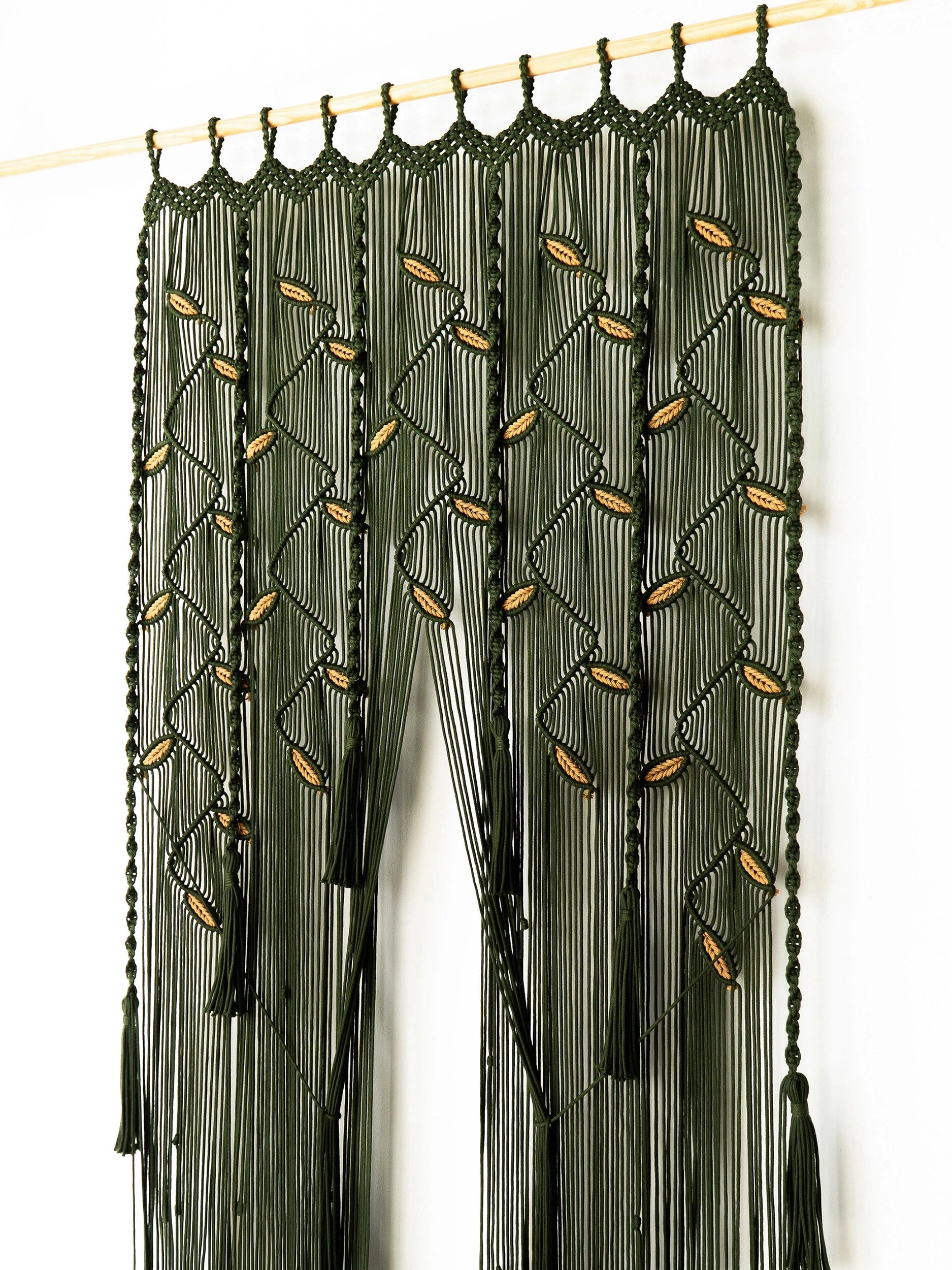 A dark olive green macrame curtain panel hanging from a light wood rod against a plain white wall. The curtain features intricate woven geometric patterns with small tan leaf-like accents and long fringe tassels.