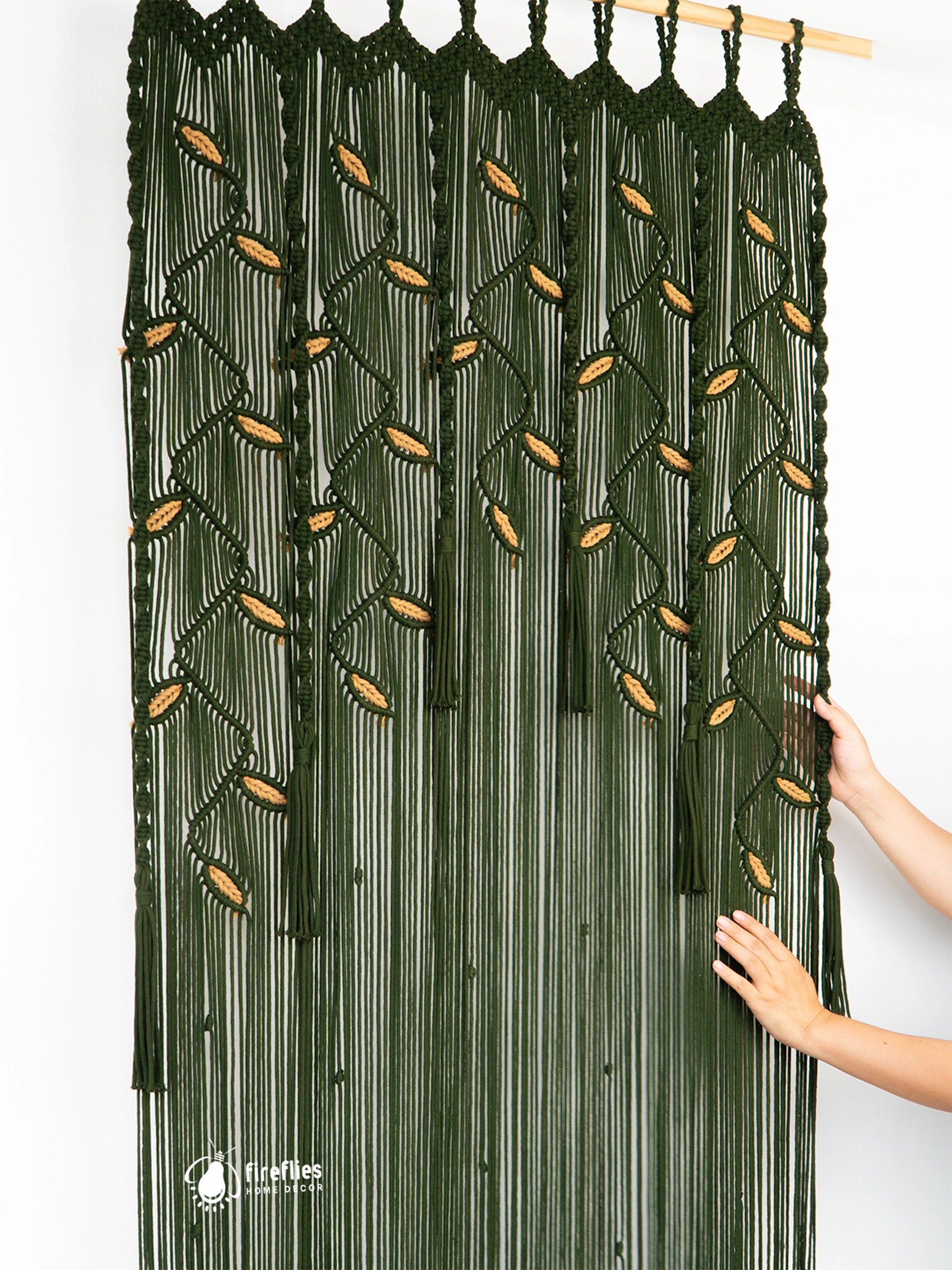 A dark green macrame wall hanging with vertical fringe and woven leaf-like motifs, shown against a white wall with a person's hands adjusting the right side.