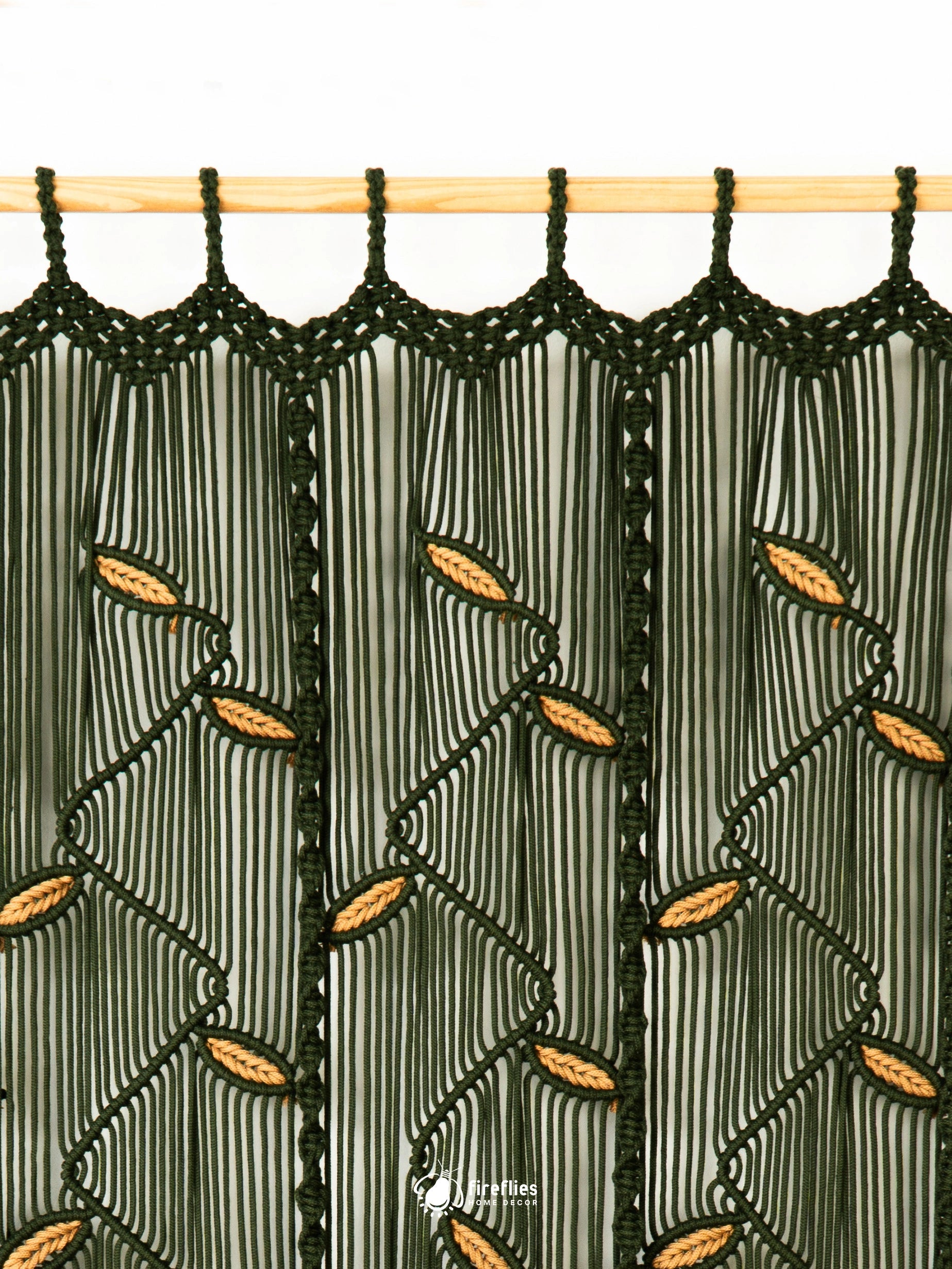 A close-up of a macrame curtain panel hanging from a light wood rod. The dark green woven cord forms a geometric pattern with vertical stripes and diagonal leaf-like accents, set against a light gray background.