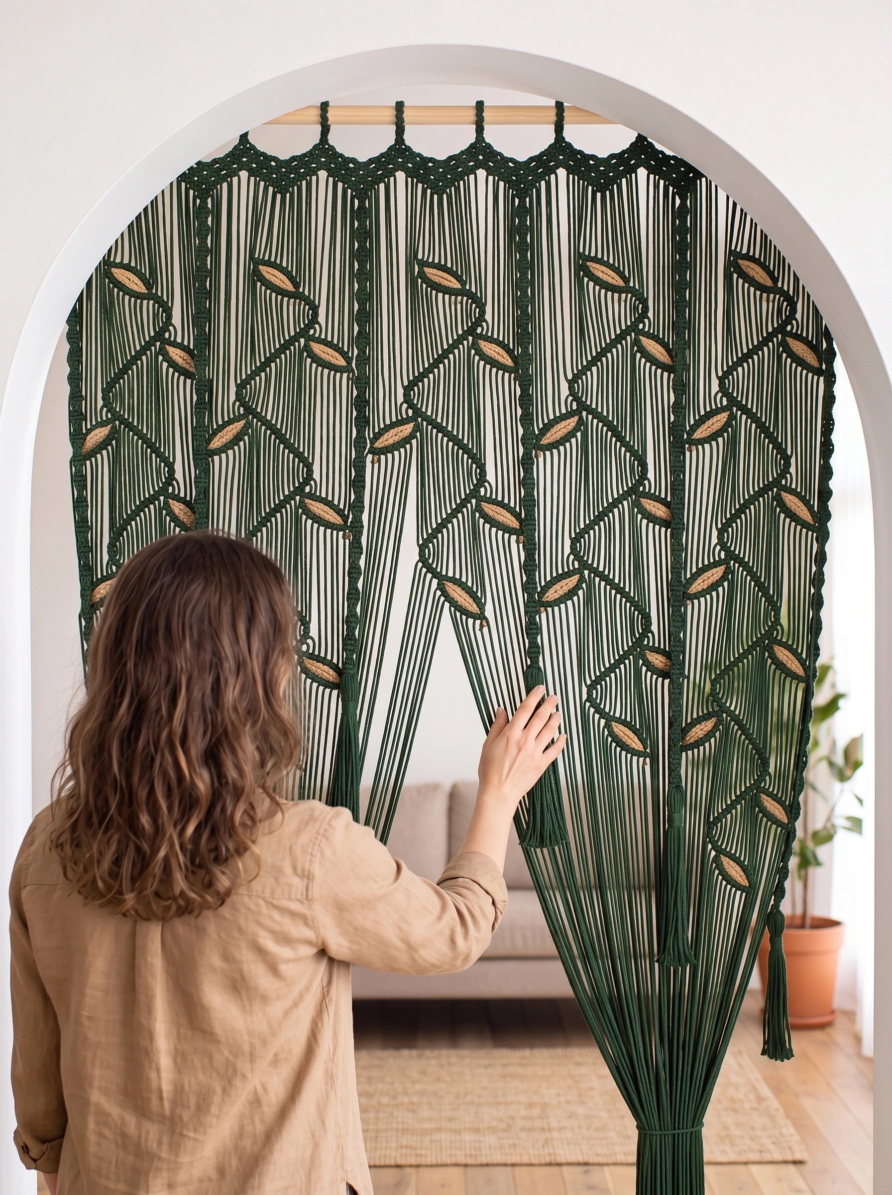A woman in a beige shirt stands in front of a large woven macrame curtain in forest green and tan, holding it open in a bright room with an arched doorway.