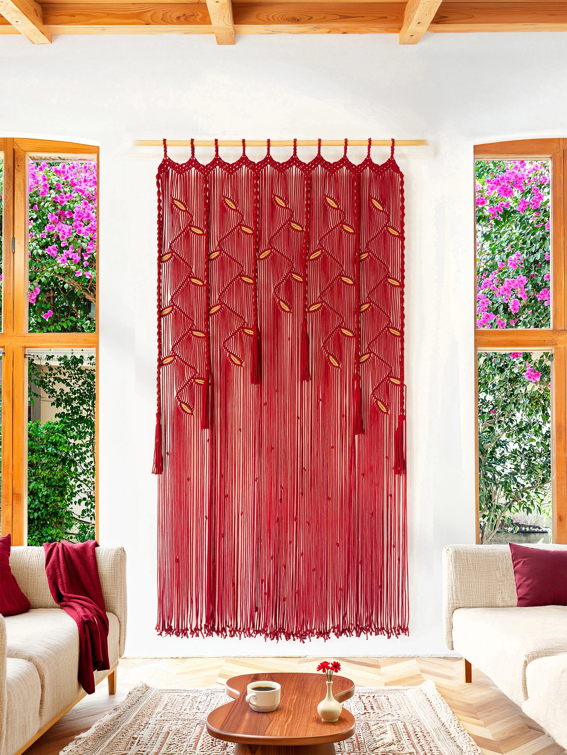 A long red sheer curtain panel with embroidered leaf motifs and tassel ties, hanging on a wooden rod in a bright living room with white walls, wood ceiling beams, and windows showing greenery outside.