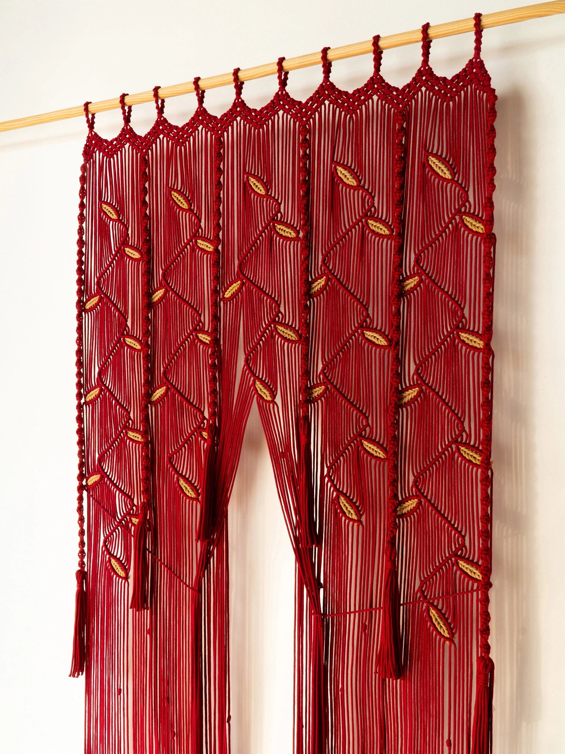 A red beaded curtain hanging from a light wood rod against a plain white wall. The curtain features intricate beadwork with leaf-like patterns in red and gold, and long dangling strands that create a tunnel effect.