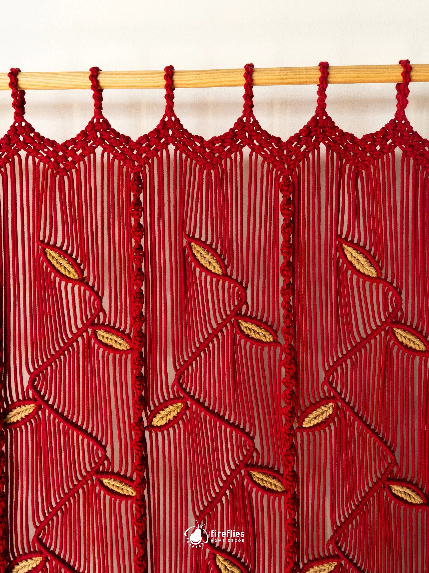 A red macrame curtain panel hanging from a light wood rod, featuring vertical fringe and woven leaf patterns with small gold accents.