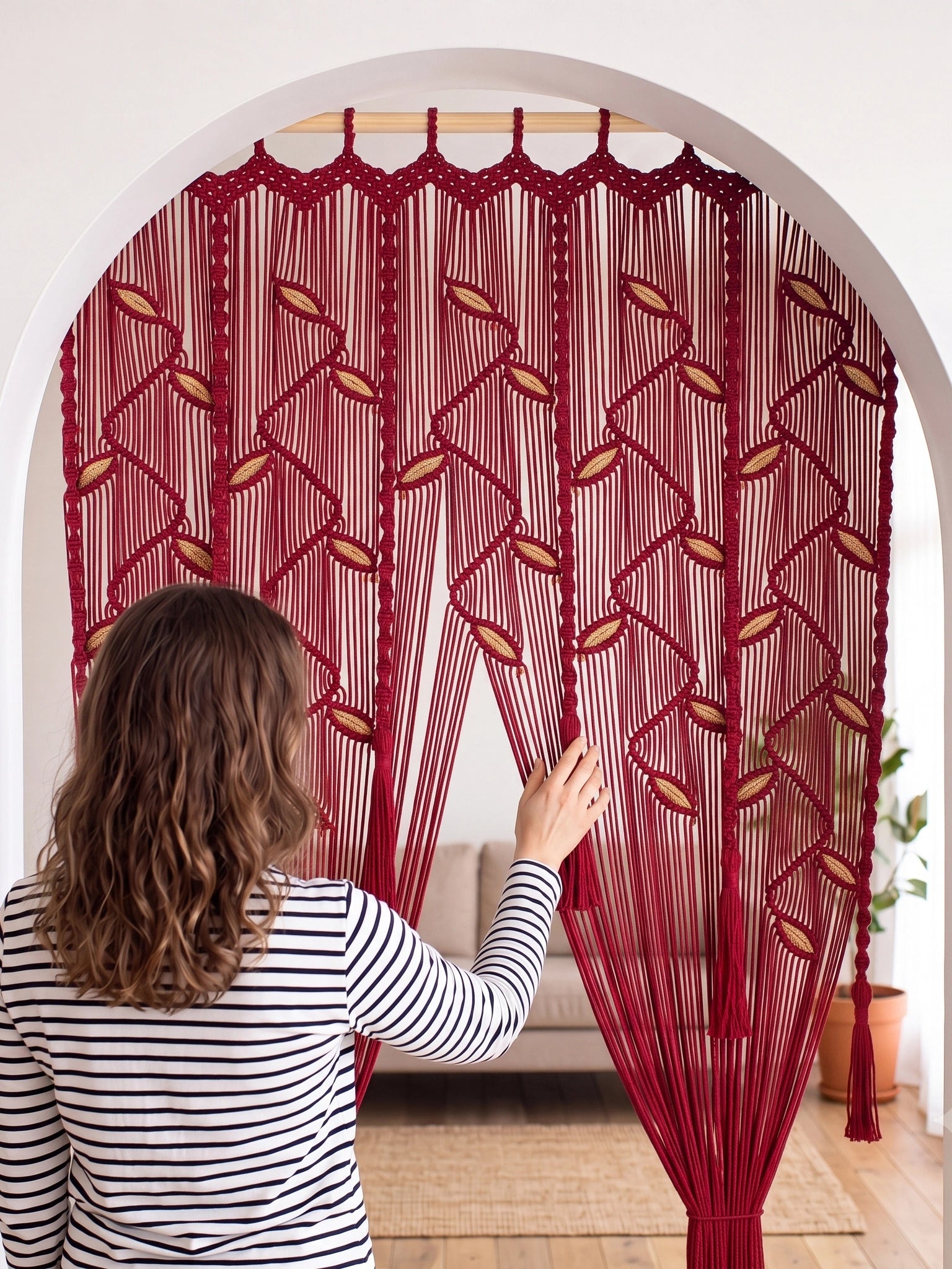 A woman in a black and white striped shirt stands in front of a red macrame curtain with leaf patterns, holding it open inside a home setting.