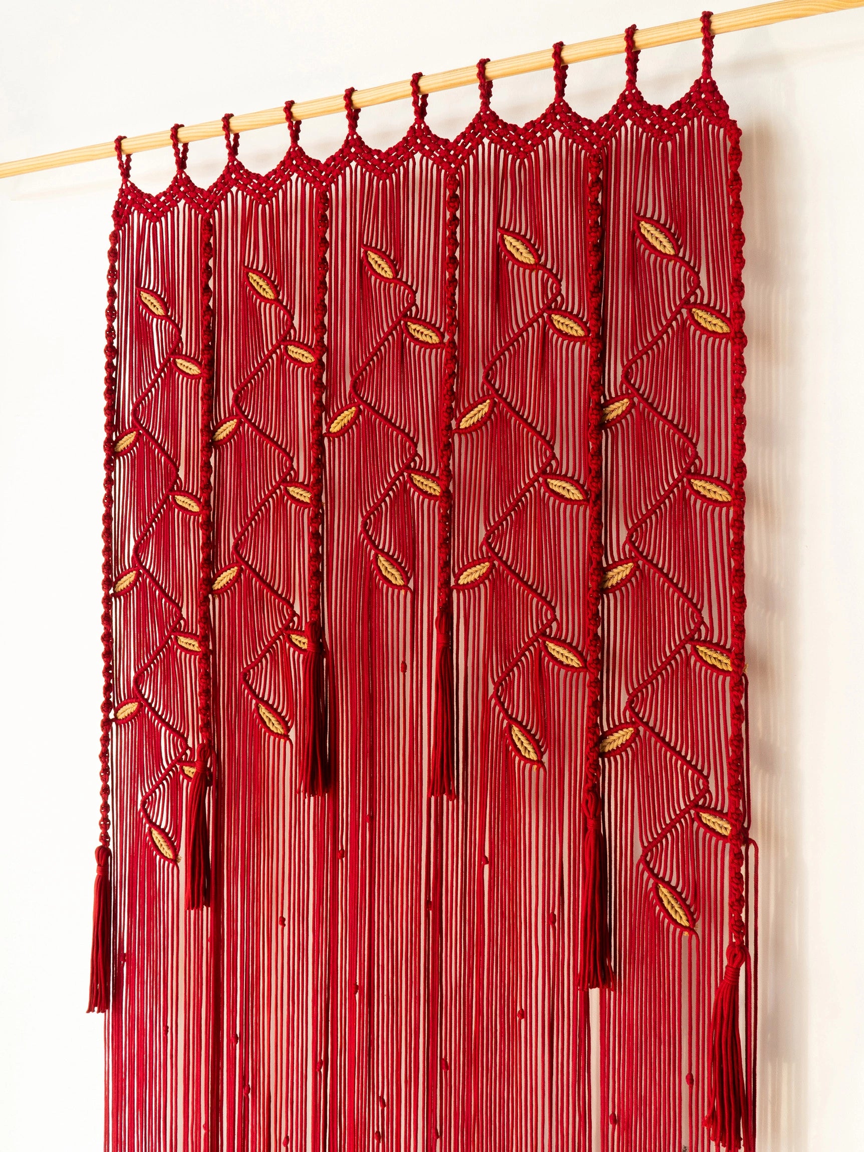 A red macrame wall hanging curtain with intricate woven patterns, hanging from a light wood rod against a plain white wall.