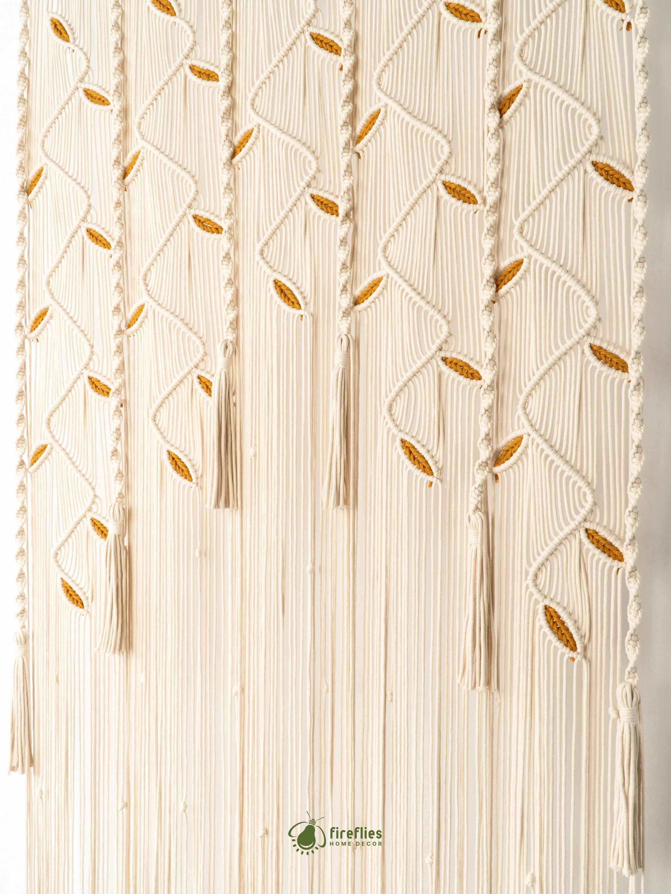 A cream-colored macrame wall hanging with intricate woven geometric patterns and small gold leaf accents, shown against a plain white background.